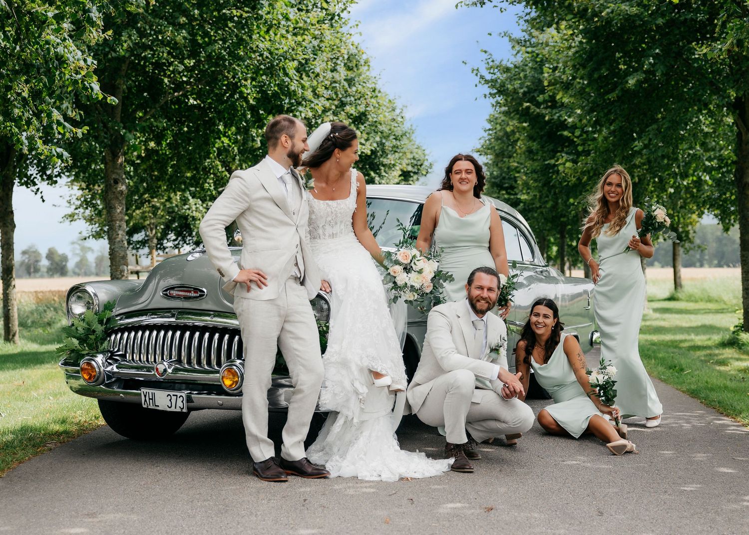 Wedding party poses playfully with vintage car on tree-lined road.