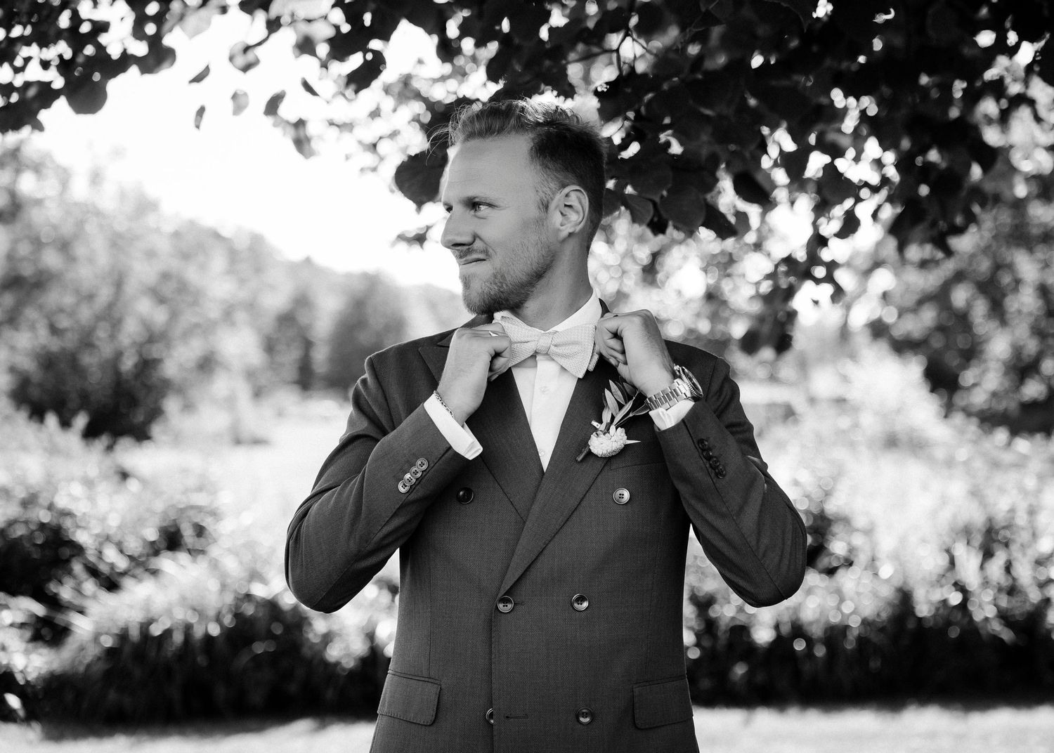 Groomsmen in black suits standing outdoors under trees in black and white photography.