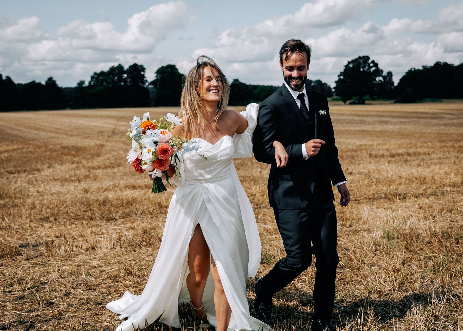 Wedding couple resting in harvested field under cloudy sky.