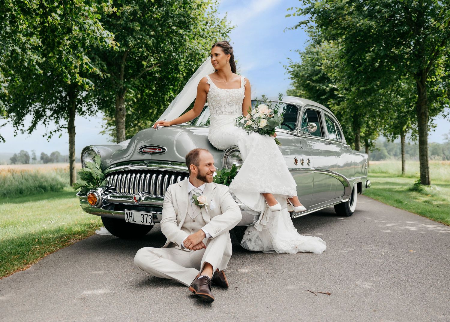 Newlyweds strike romantic poses with vintage automobile under green canopy.