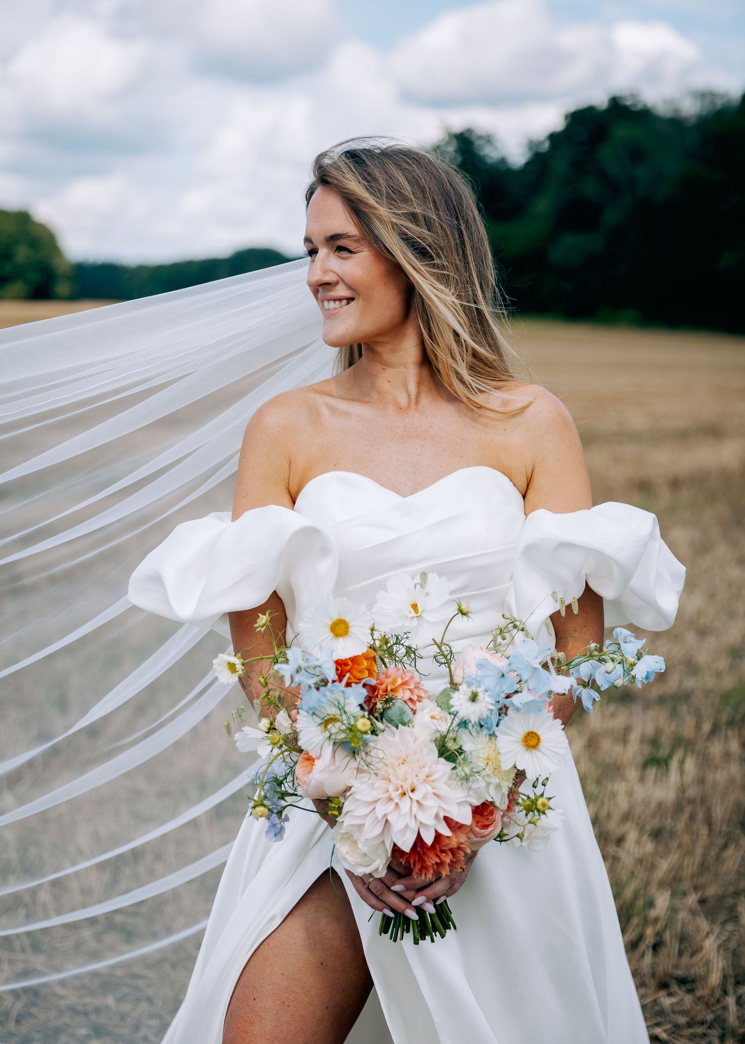 Bride with flowing veil holds colorful summer bouquet in wheat field.