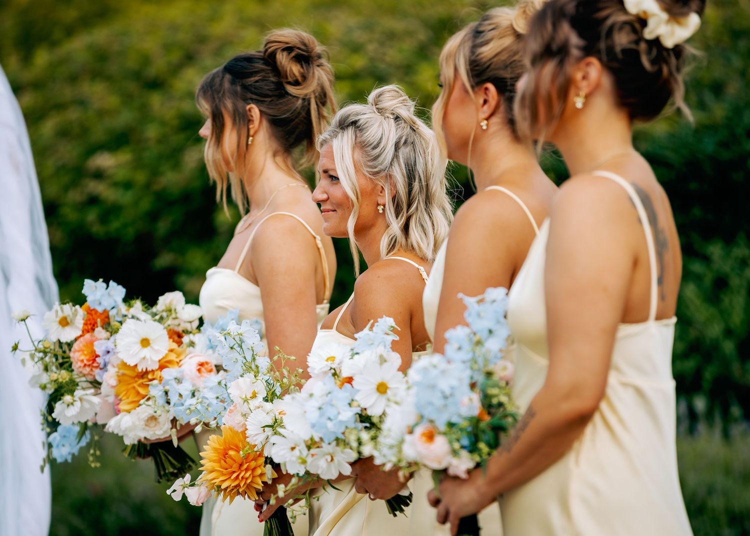 Bridesmaids in light colored dresses hold vibrant orange and white floral bouquets at an outdoor wedding ceremony.