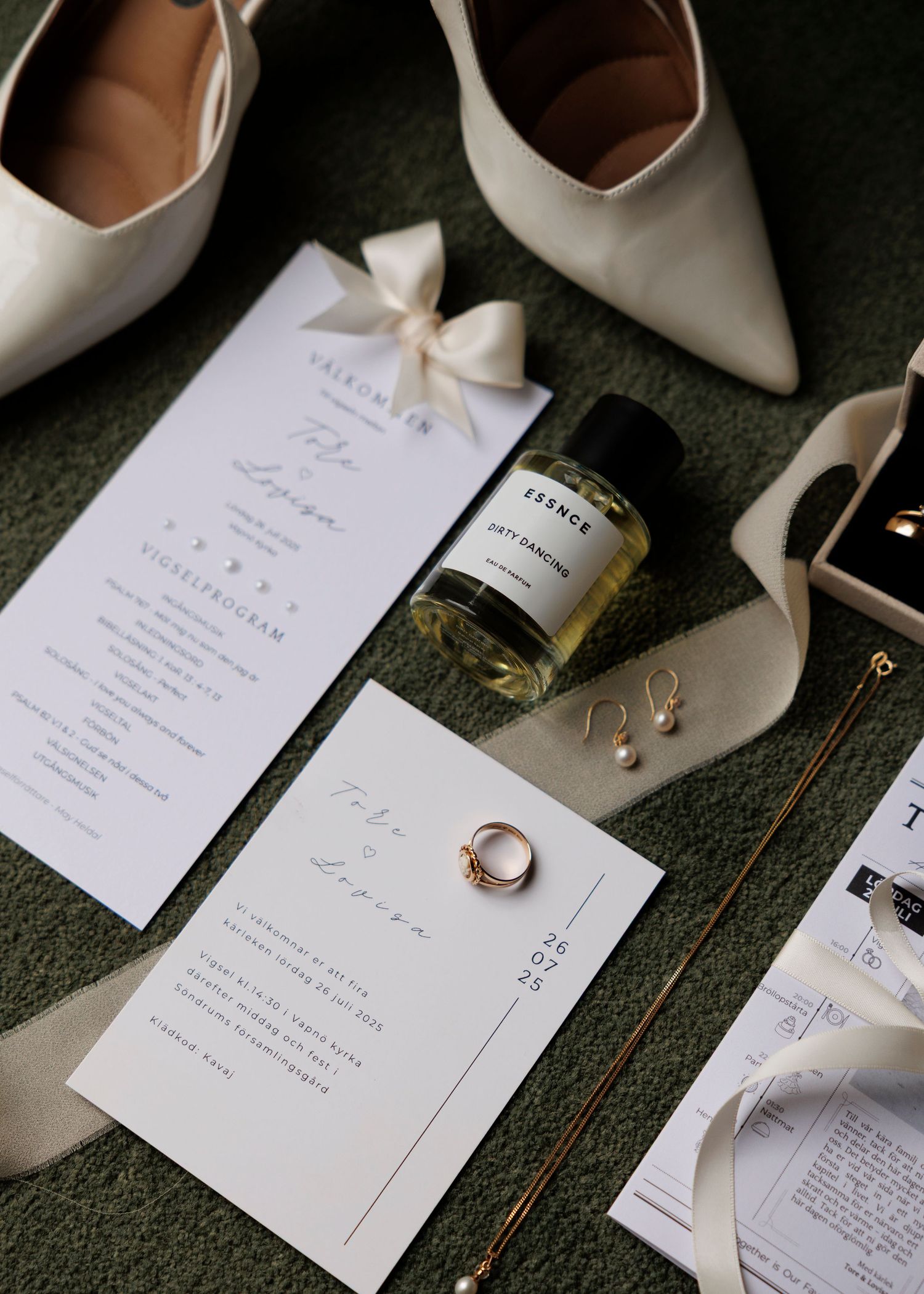 Wedding details flatlay with white shoes, invitations and rings on dark surface.