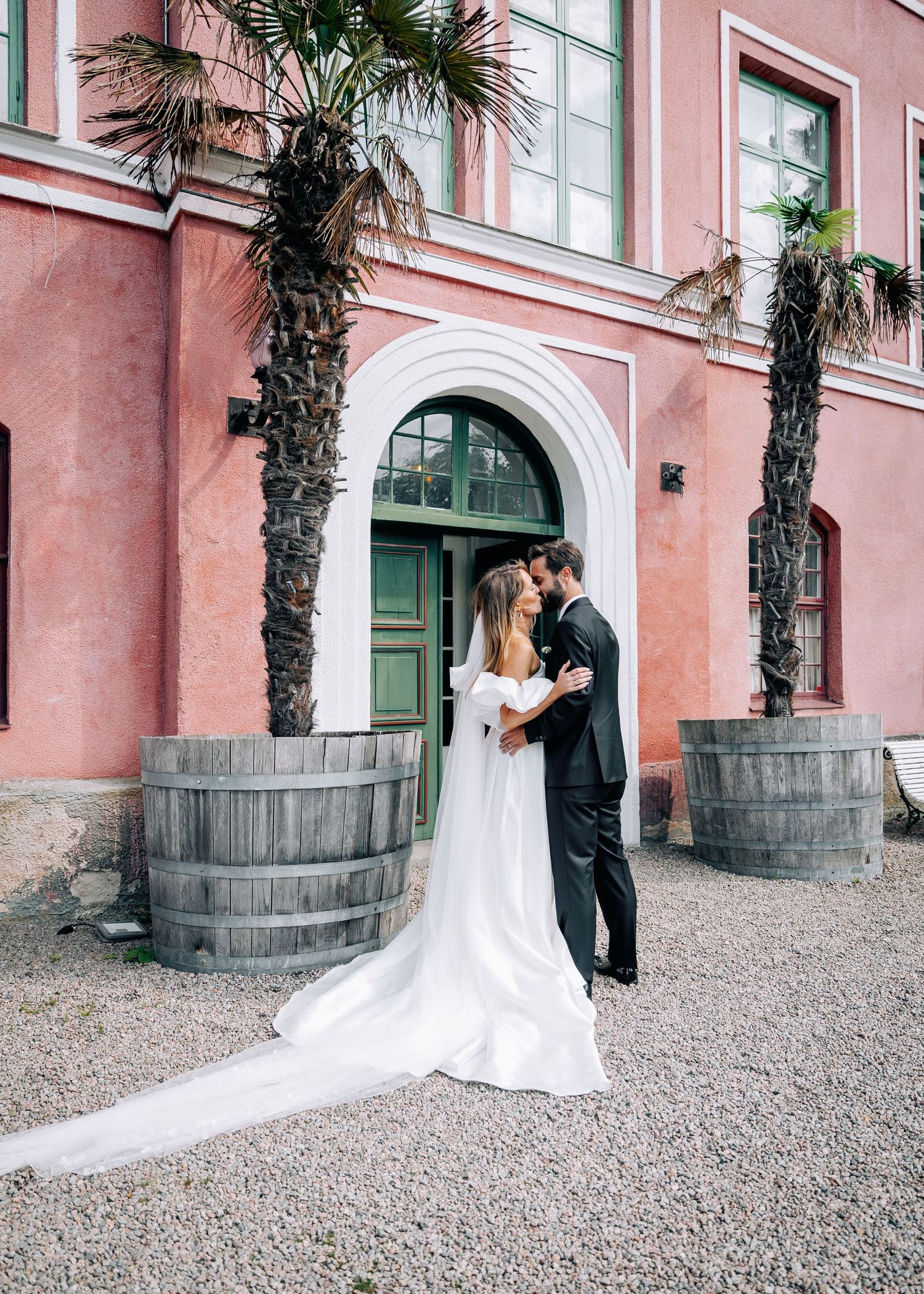 Wedding couple poses against pink stucco building with palm trees creating elegant architectural backdrop.