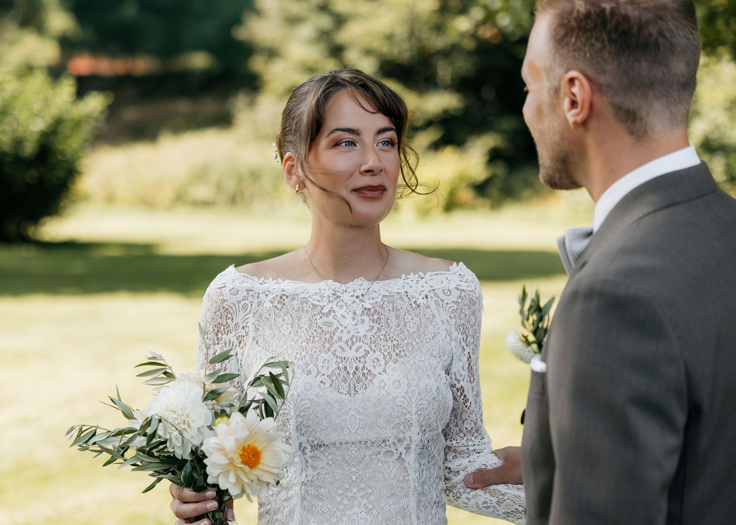 Bride in lace dress and groom in dark suit share an intimate moment outdoors.