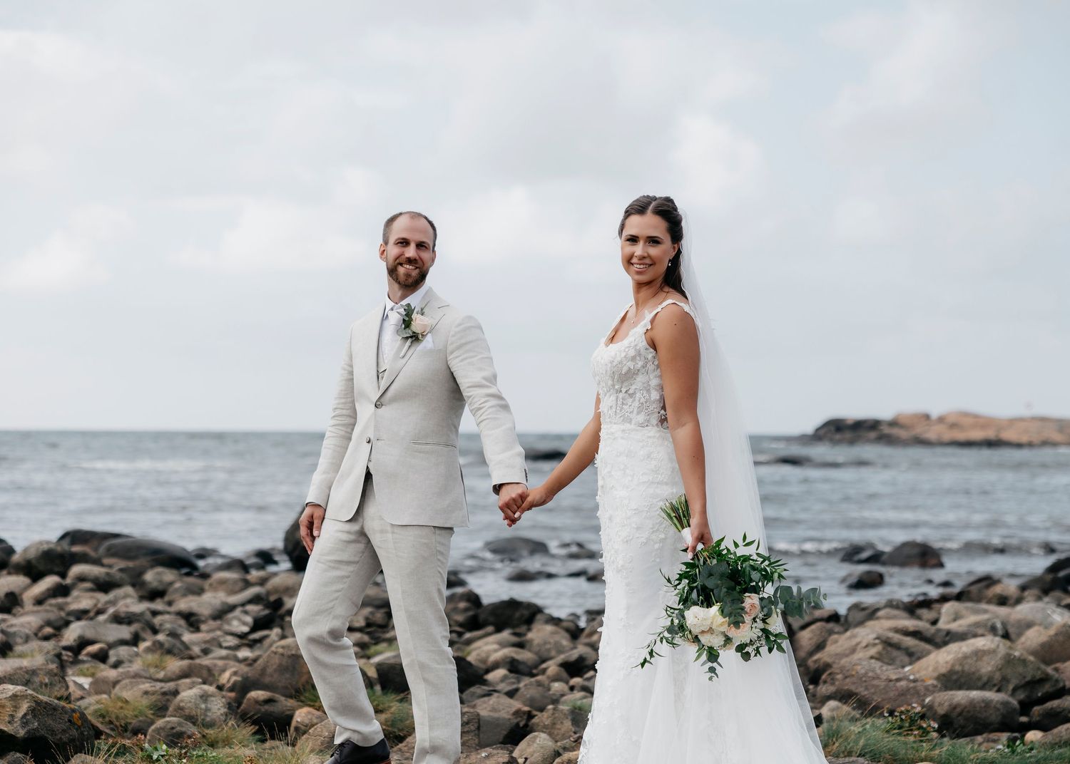 Newlyweds pose together on rocky beach with waves and coastline in background.
