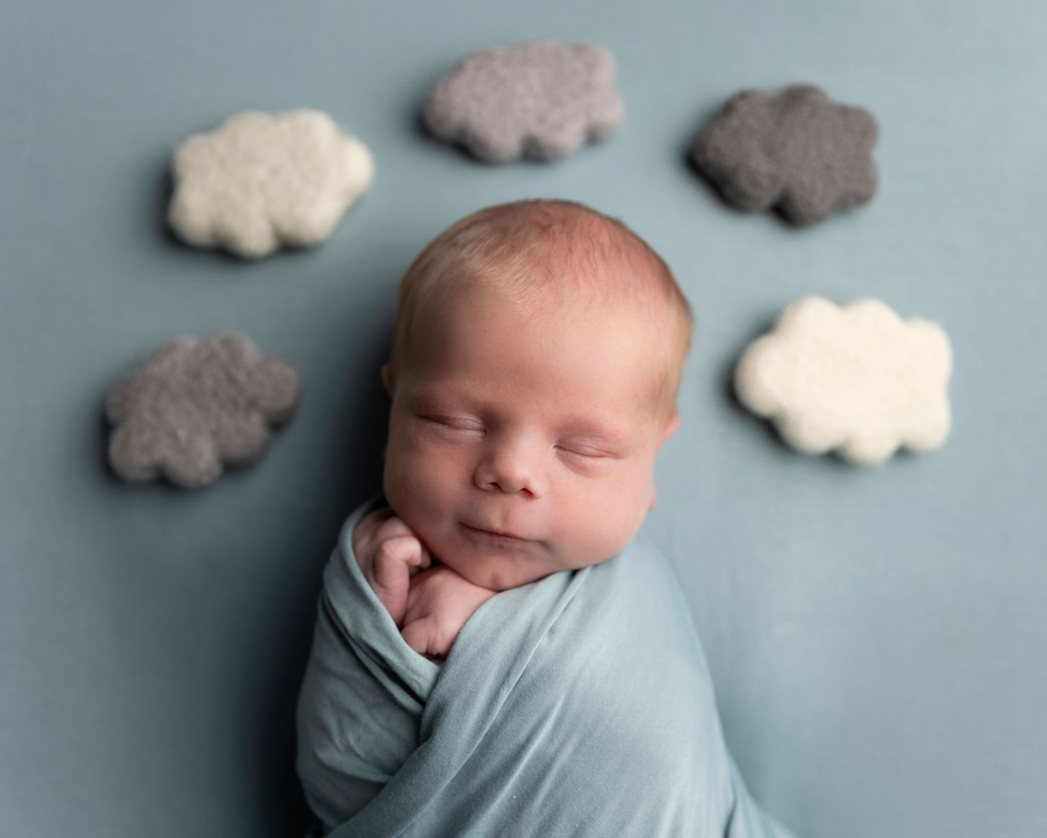 Sleeping newborn wrapped in light blue swaddle with decorative cloud shapes in the background.