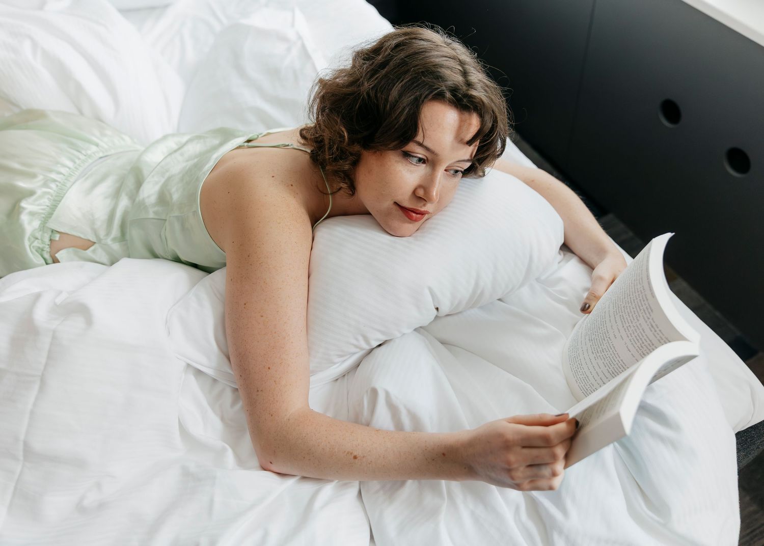 Person relaxing in white bedding while reading in natural morning light.