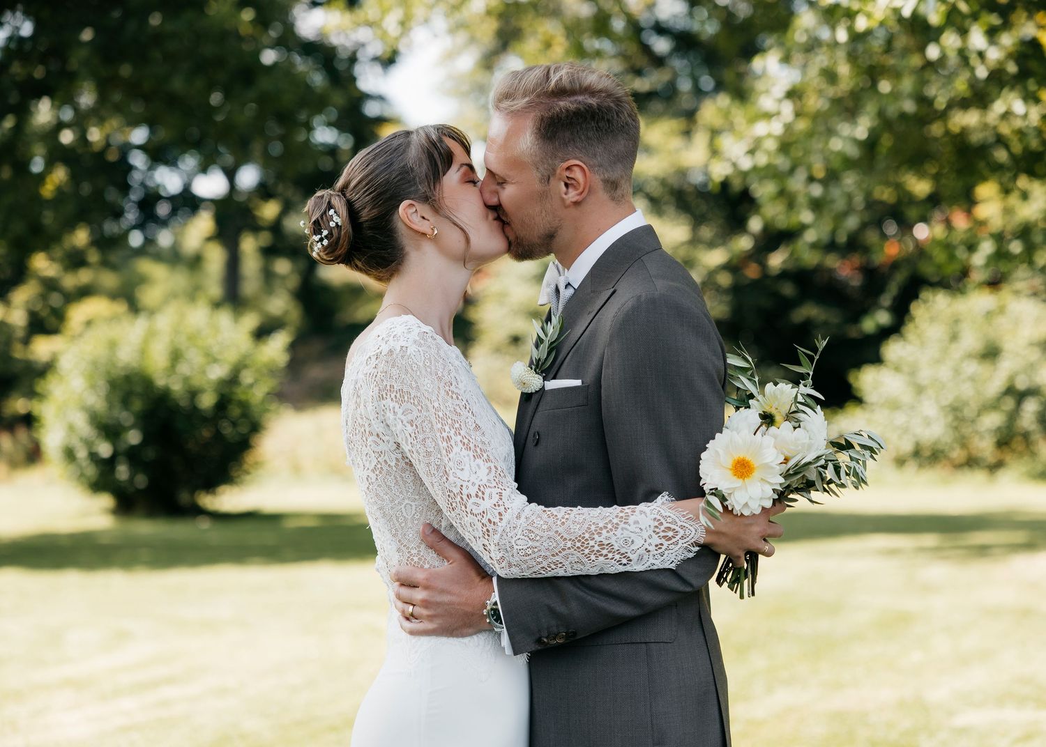A romantic wedding kiss in a sun-drenched garden with a bouquet of white and yellow flowers.