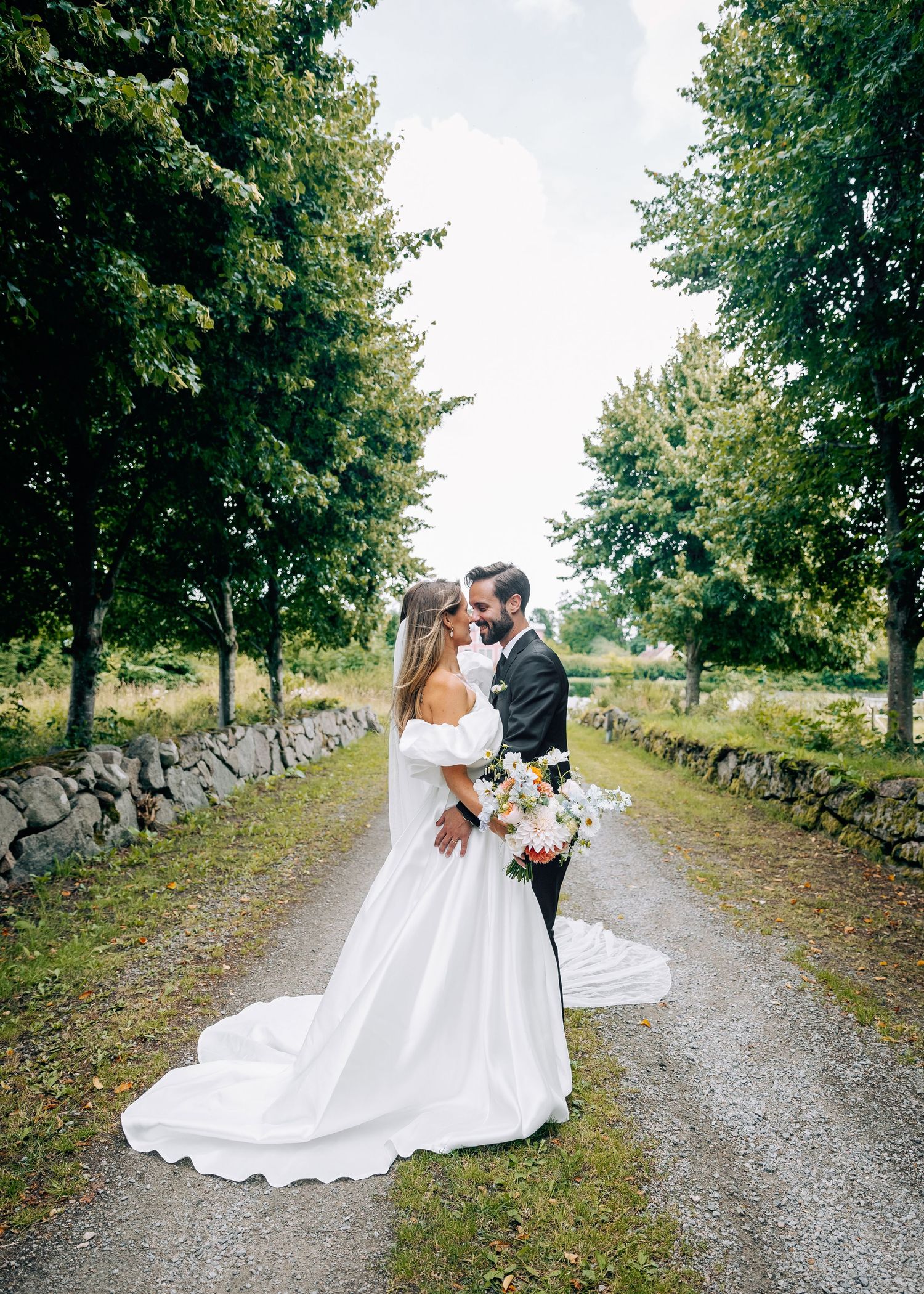 Wedding couple walks along tree-lined stone wall path in romantic countryside setting.