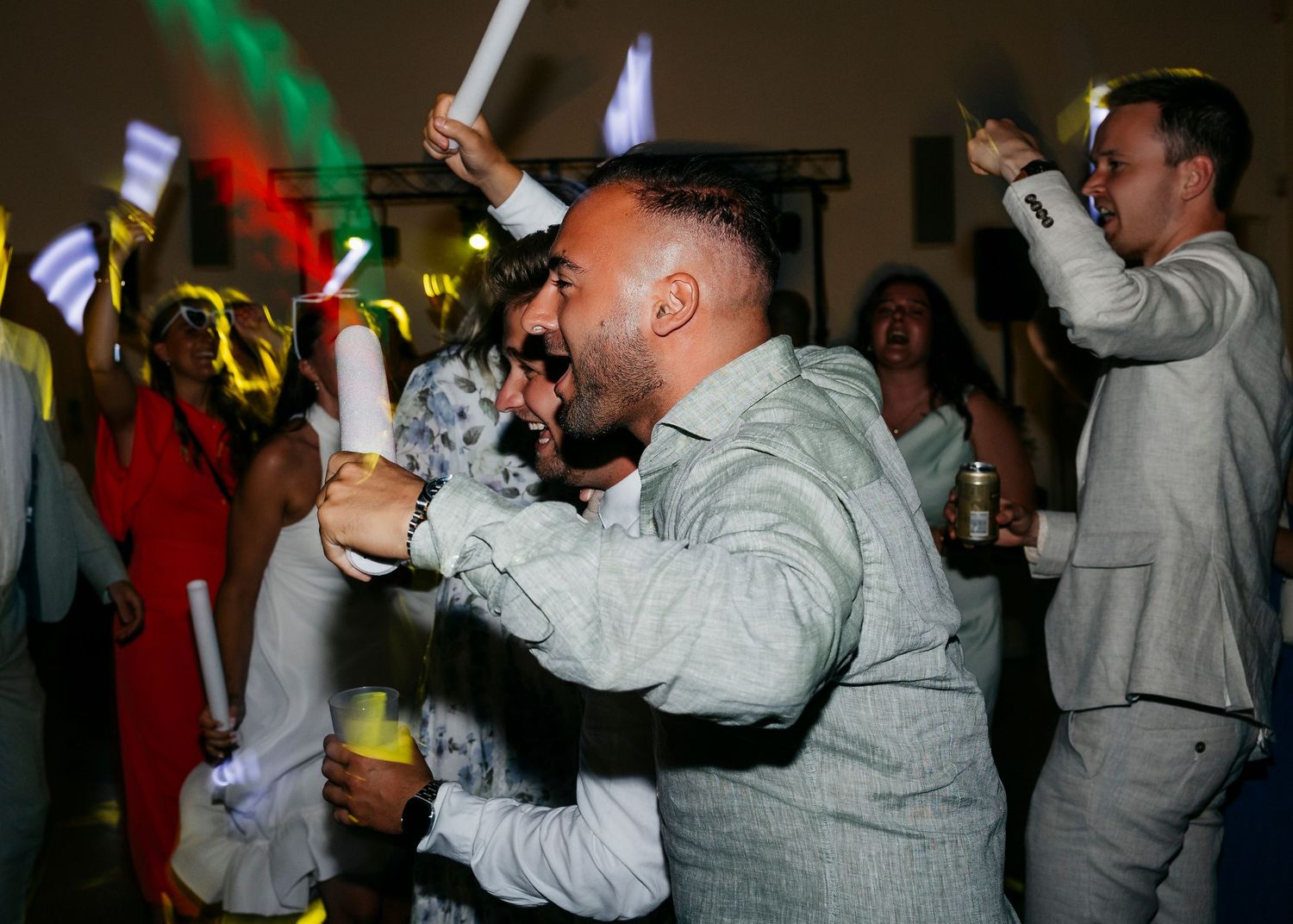 Wedding guests celebrate enthusiastically with light sticks on a colorful dance floor.