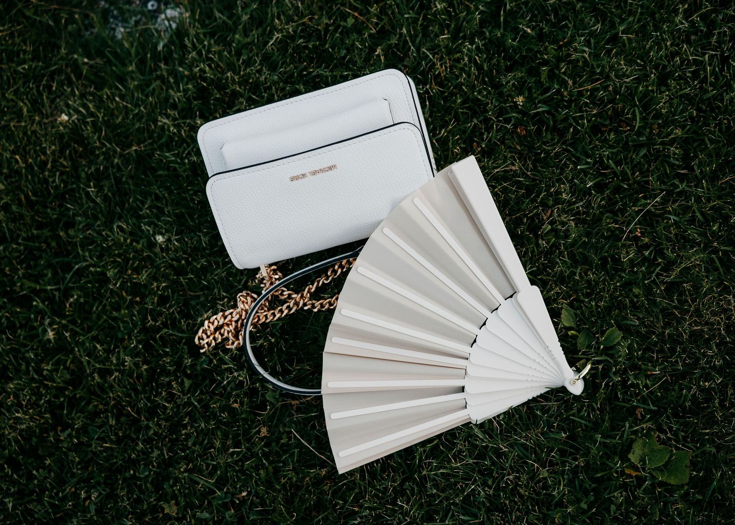 White decorative hand fan and small purse displayed on grass.