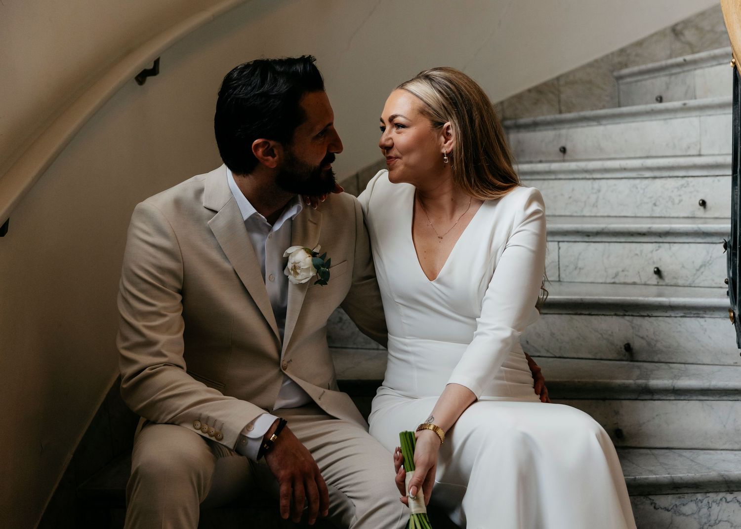 A couple shares romantic moments while sitting on marble stairs in matching light-colored outfits.