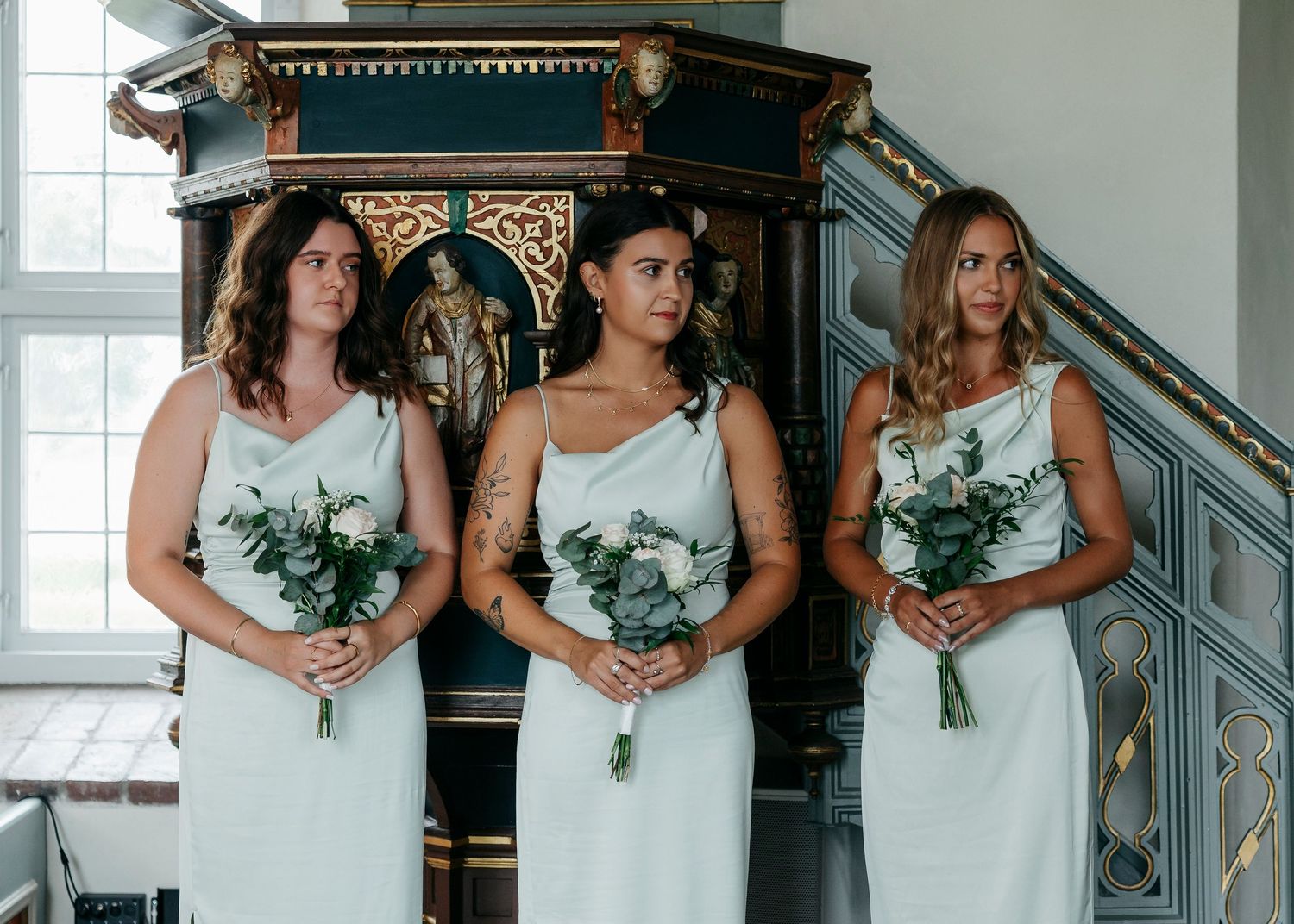 Row of bridesmaids in matching dresses holding white floral bouquets in church setting.