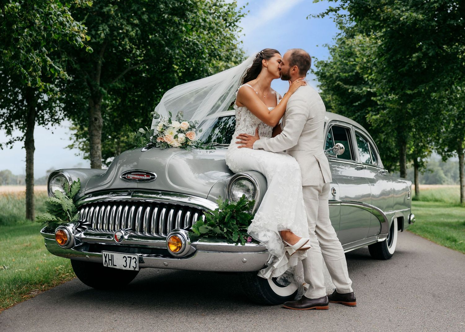 Elegant wedding photos featuring classic car on scenic tree-lined avenue.