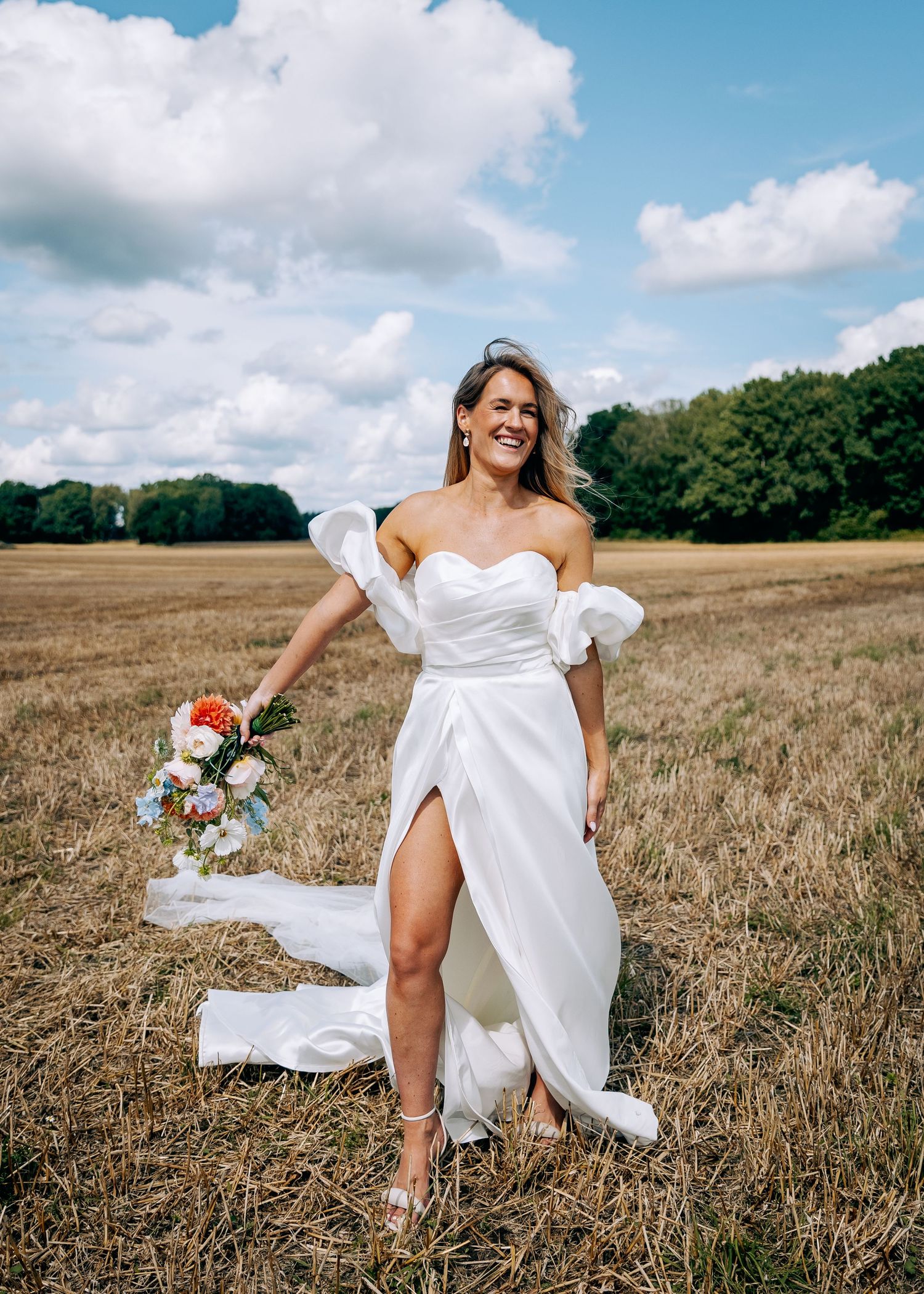 Bride in off-shoulder dress poses playfully in golden wheat field.