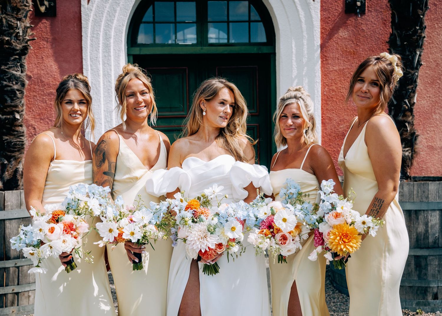 Bridal party in white dresses holding colorful bouquets pose in front of pink building.