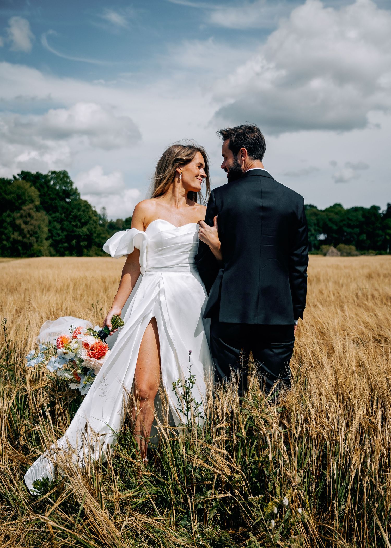 Newlywed couple walking through golden wheat field on cloudy summer day.
