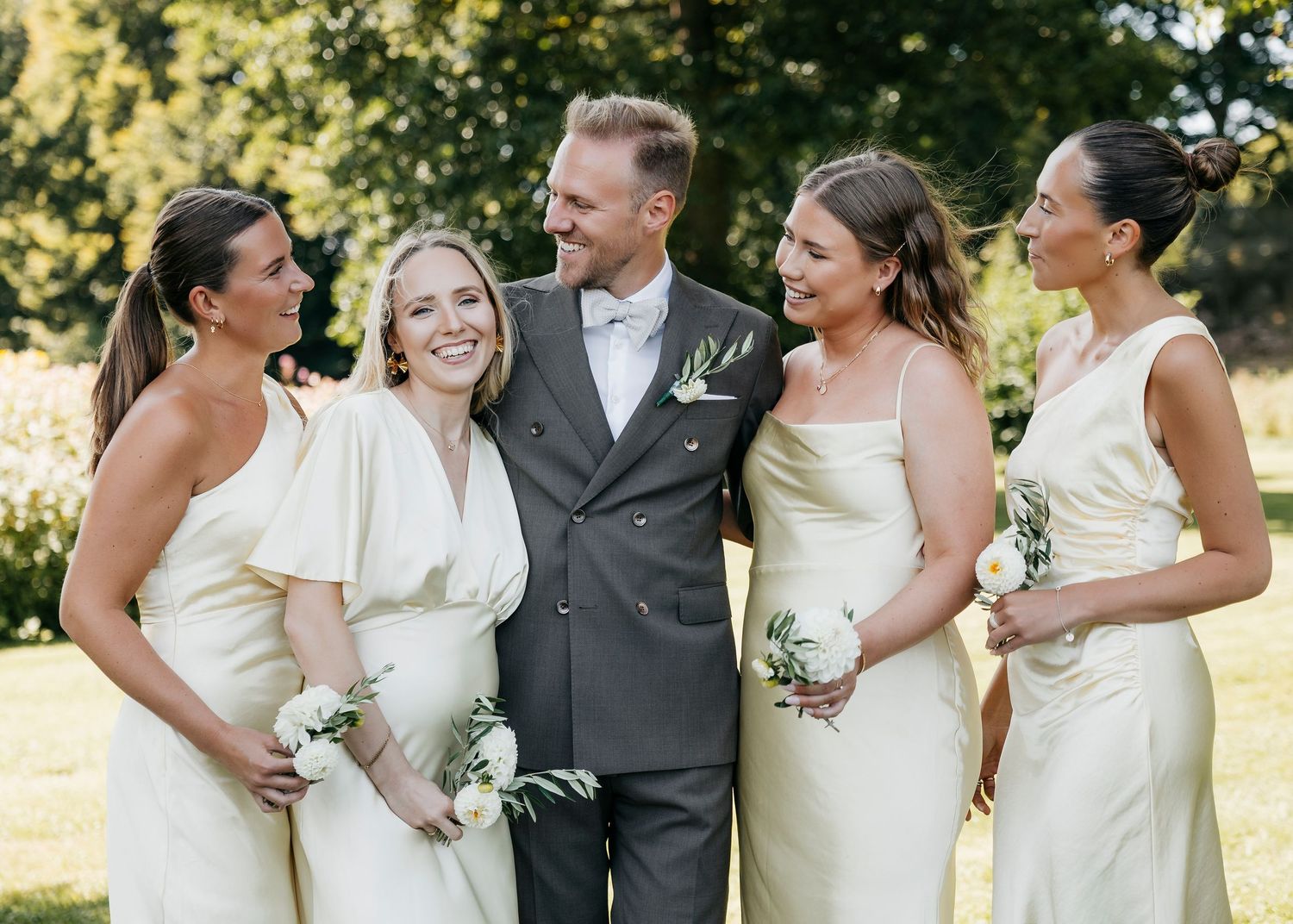 Wedding party members pose together in formal white dresses and gray attire under trees.