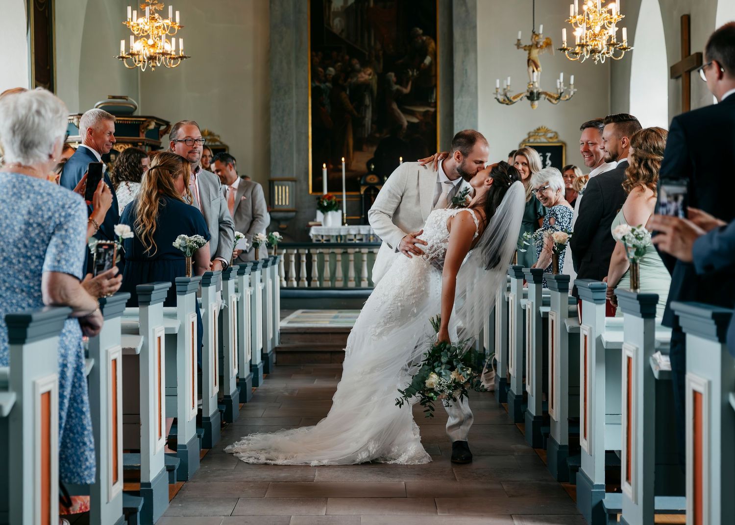 Church interior with rows of white pews and crystal chandeliers during a wedding ceremony.