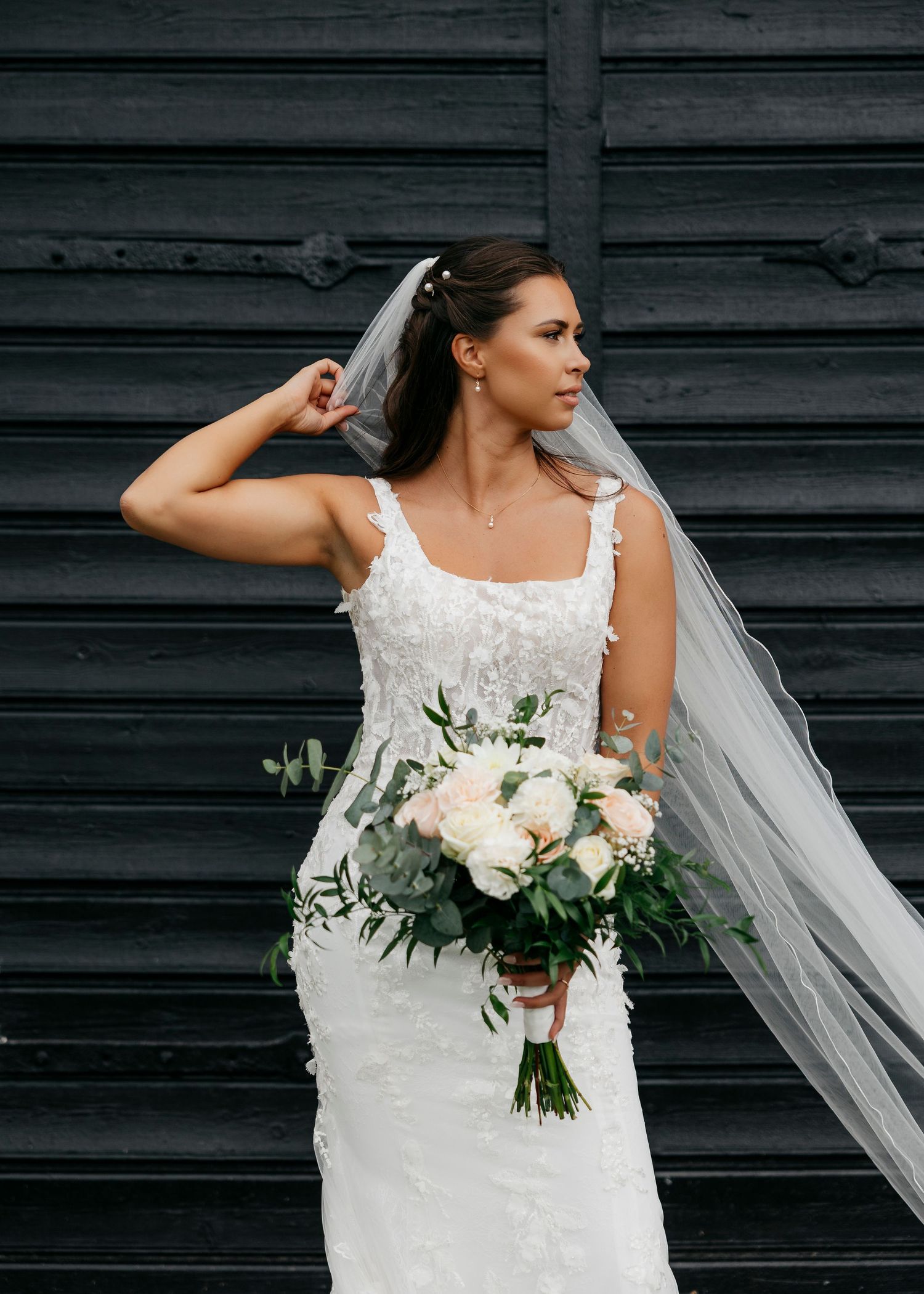 Elegant bride in white lace dress holding a romantic bouquet of white roses and greenery.