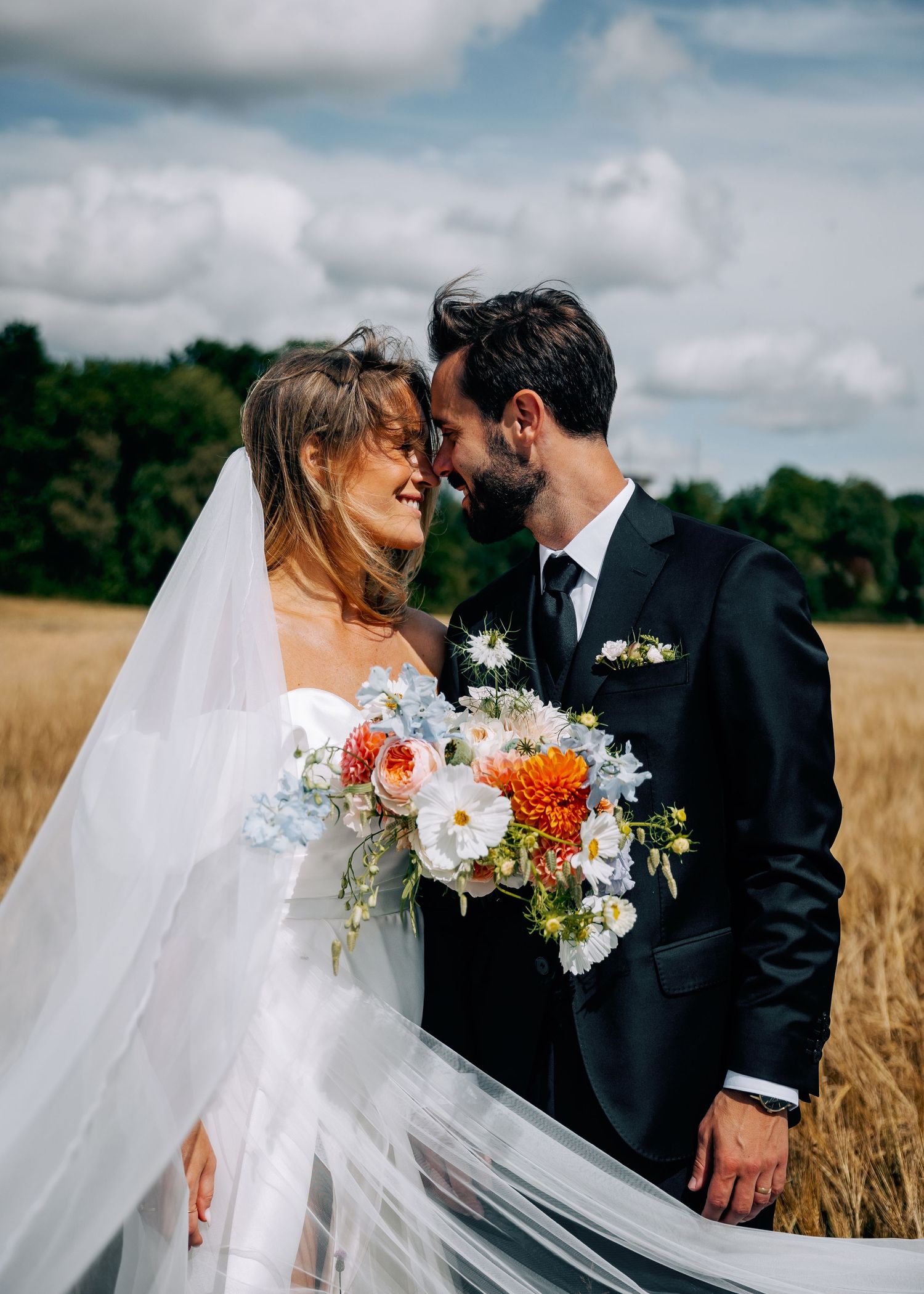 Wedding couple embracing in wheat field with flowing white veil and colorful bouquet.