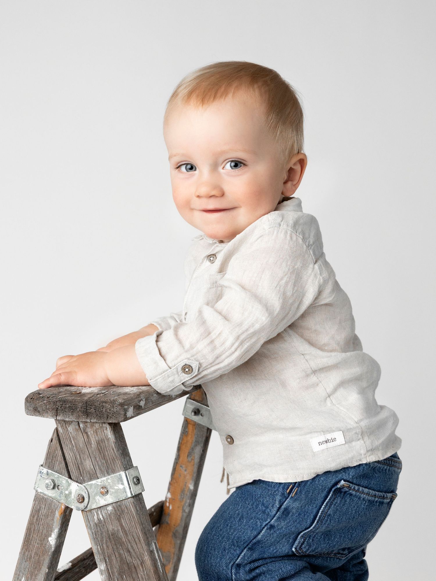 Small child in white shirt and jeans leaning on rustic wooden stool against white background.