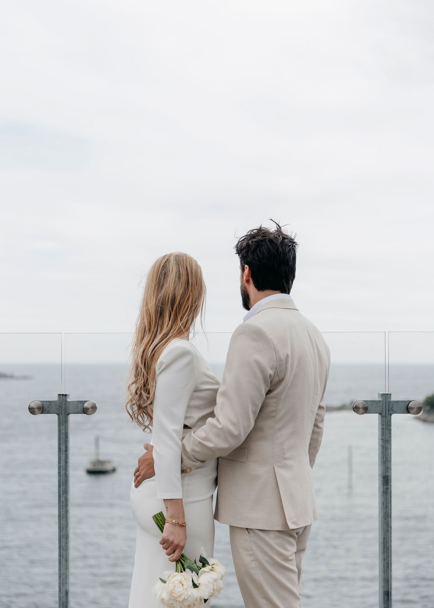 Newlywed couple in white and beige attire embracing by the ocean with a cloudy sky backdrop.