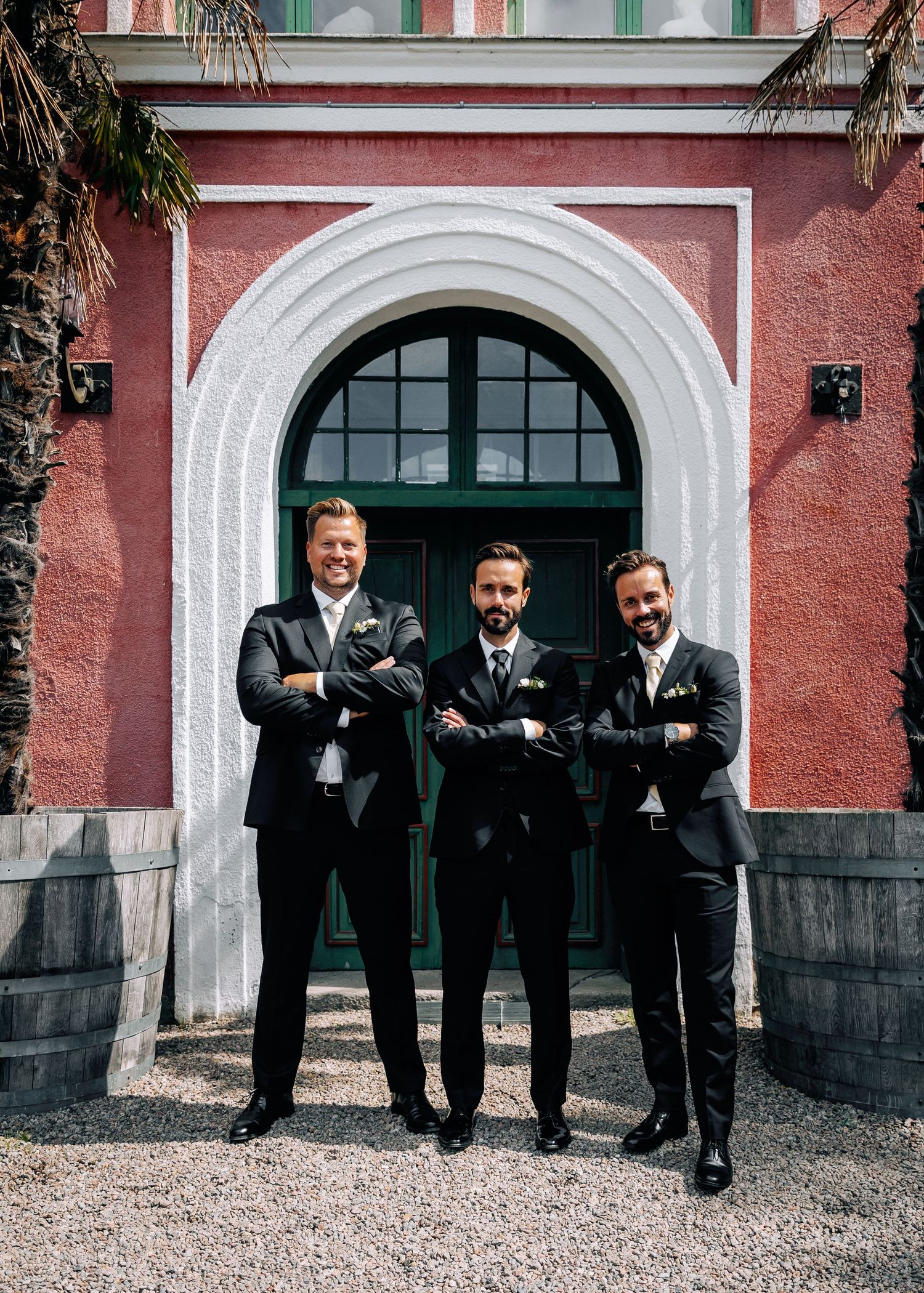 Groomsmen pose playfully against red building with white arched doorways.