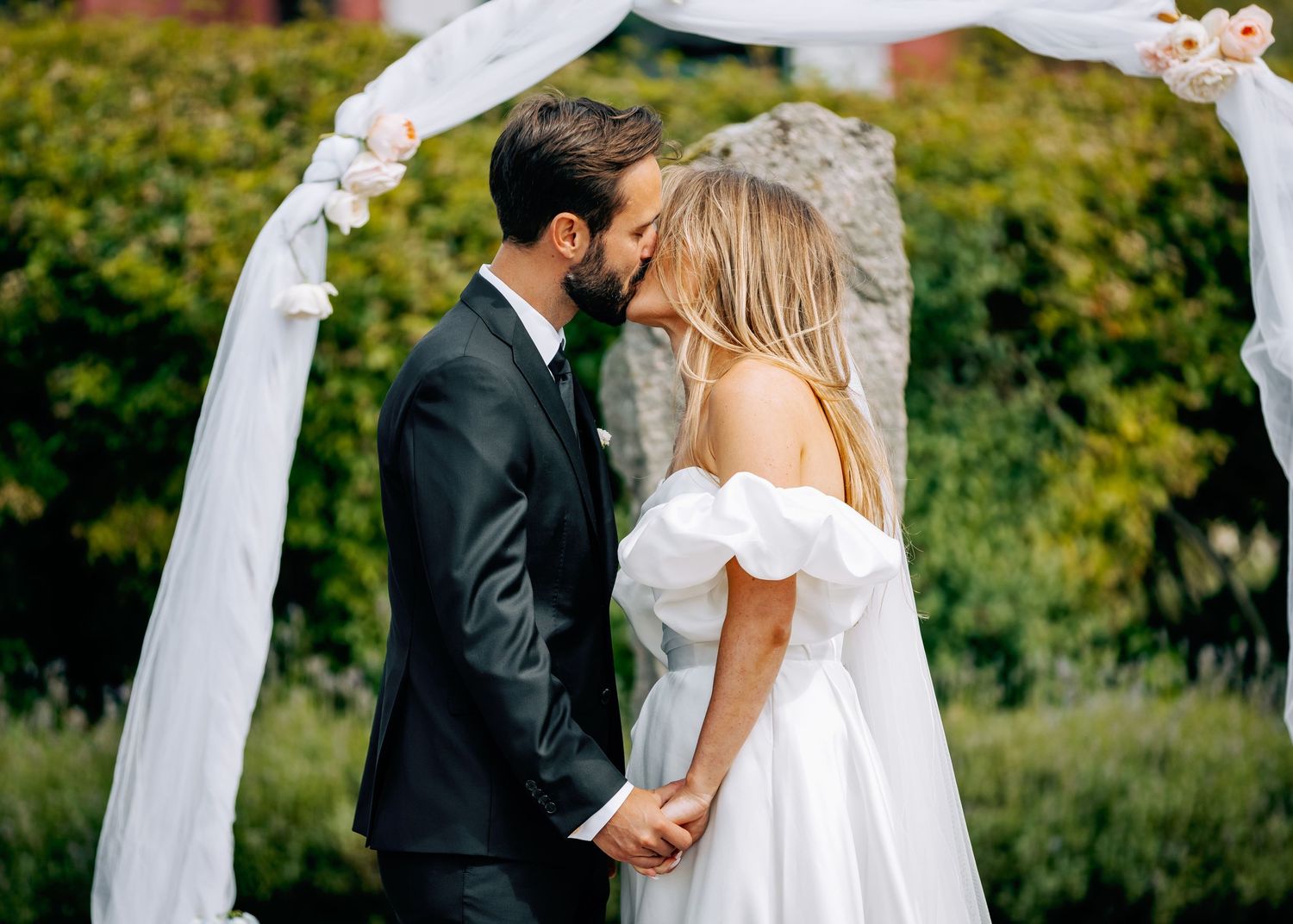 Outdoor wedding ceremony with white fabric arch and floral arrangements against pink building.