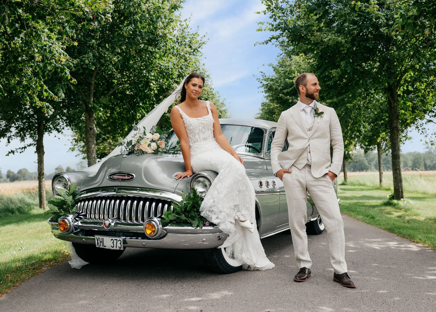 Wedding couple creates playful moments with classic car on tree-lined street.