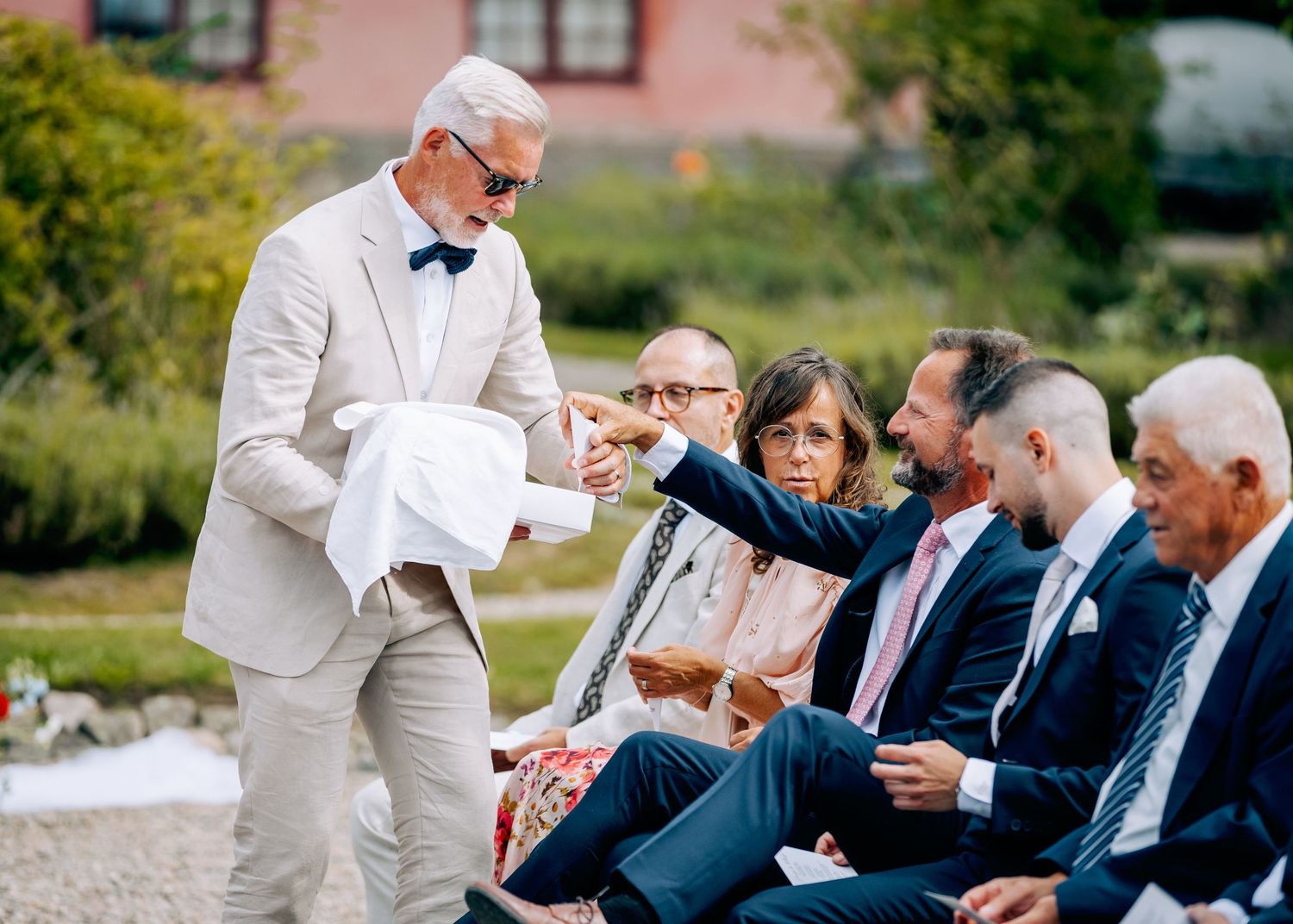 Wedding guests seated in white chairs on a gravel courtyard in front of a pink building.