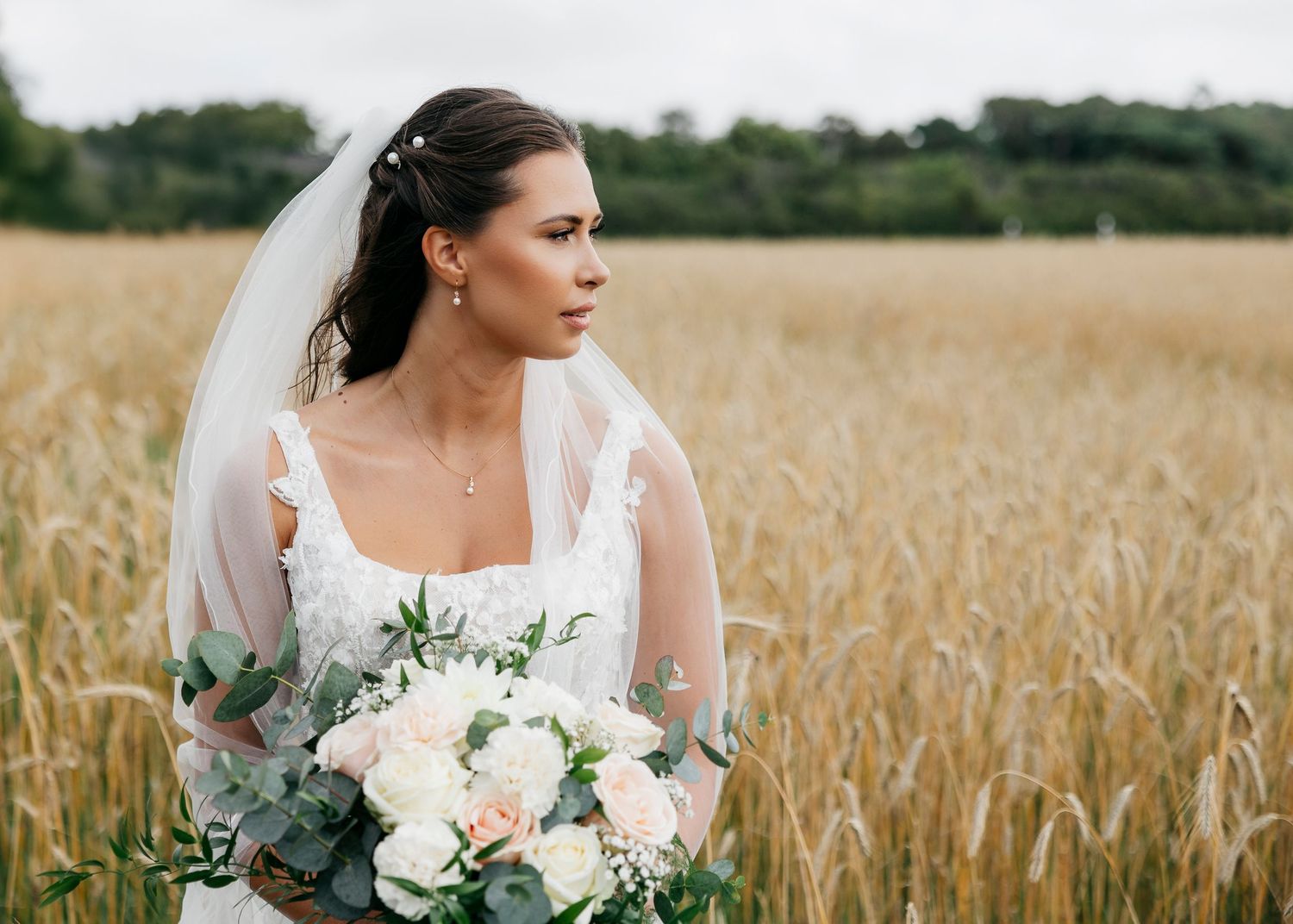 Bride holds white and green floral bouquet while standing in golden wheat field.