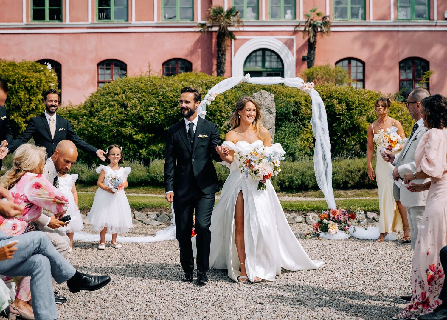 Wedding ceremony procession on gravel courtyard with pink building and white arch decorations.