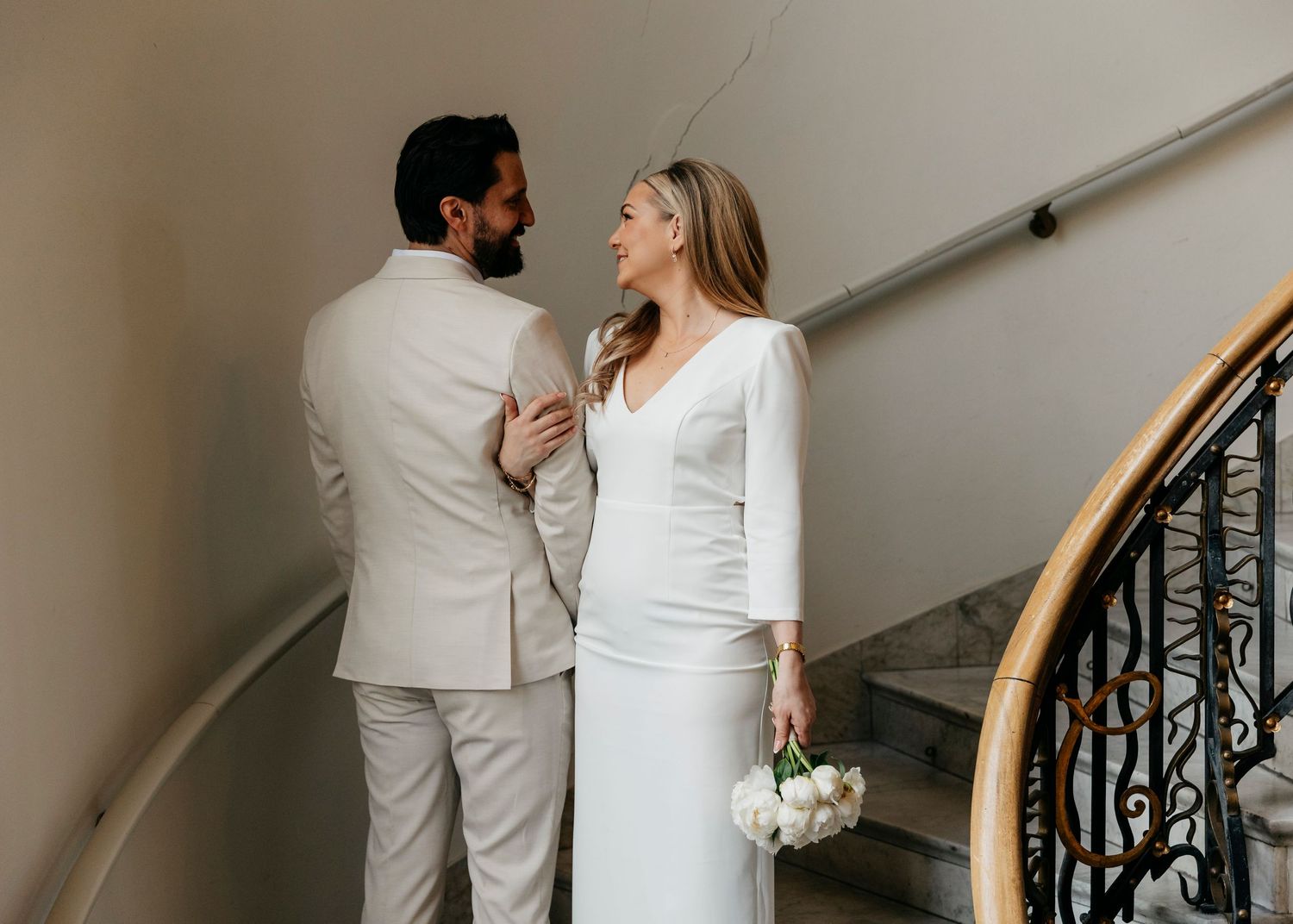 A couple in white outfits poses together on elegant marble stairs with an ornate railing.