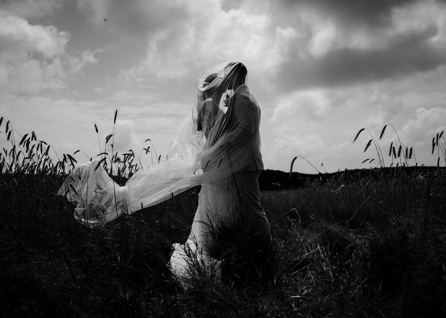 Dramatic black and white wedding photo in field with flowing veil against stormy sky.