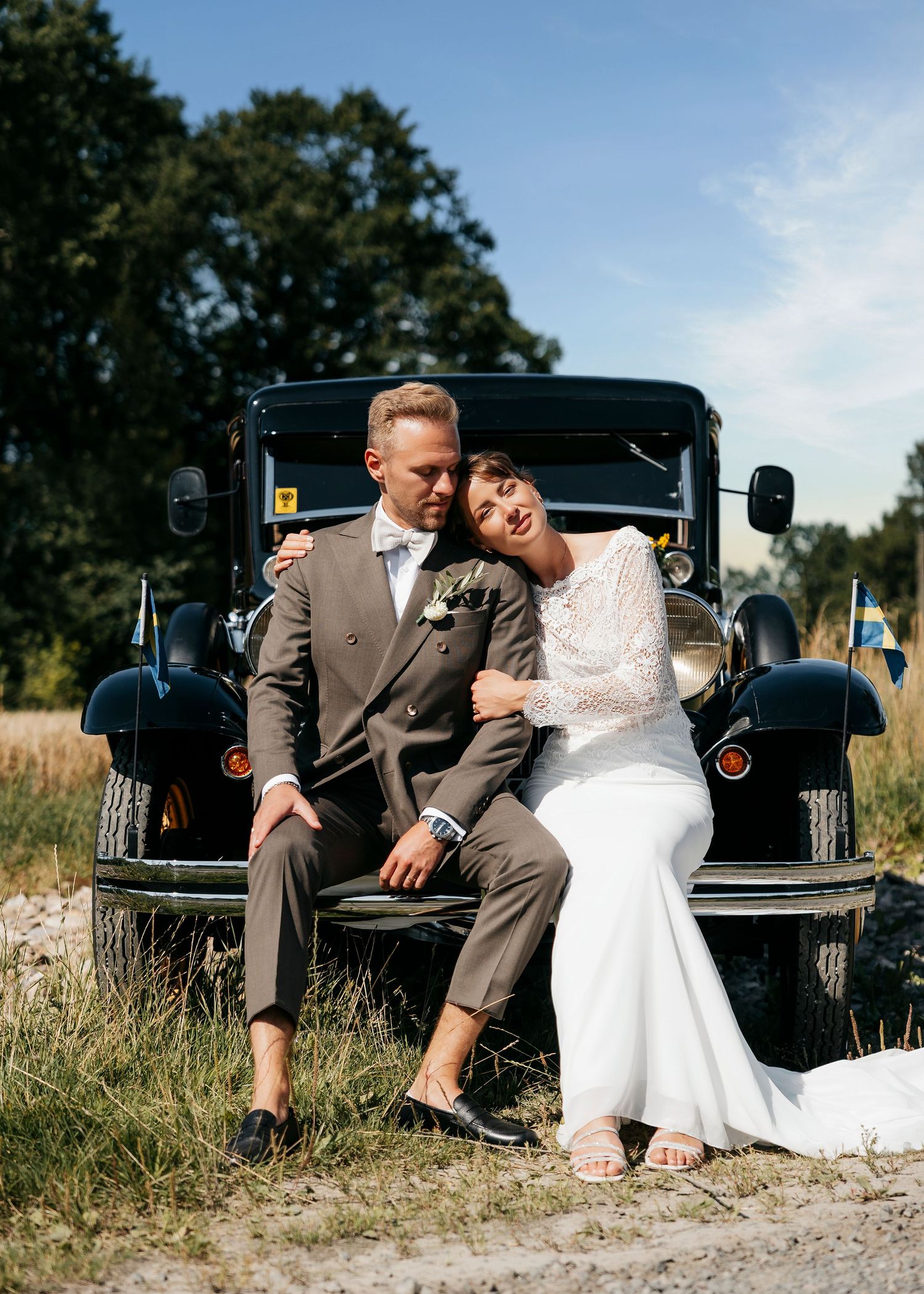 Wedding couple relaxes on vintage car in rustic outdoor setting.