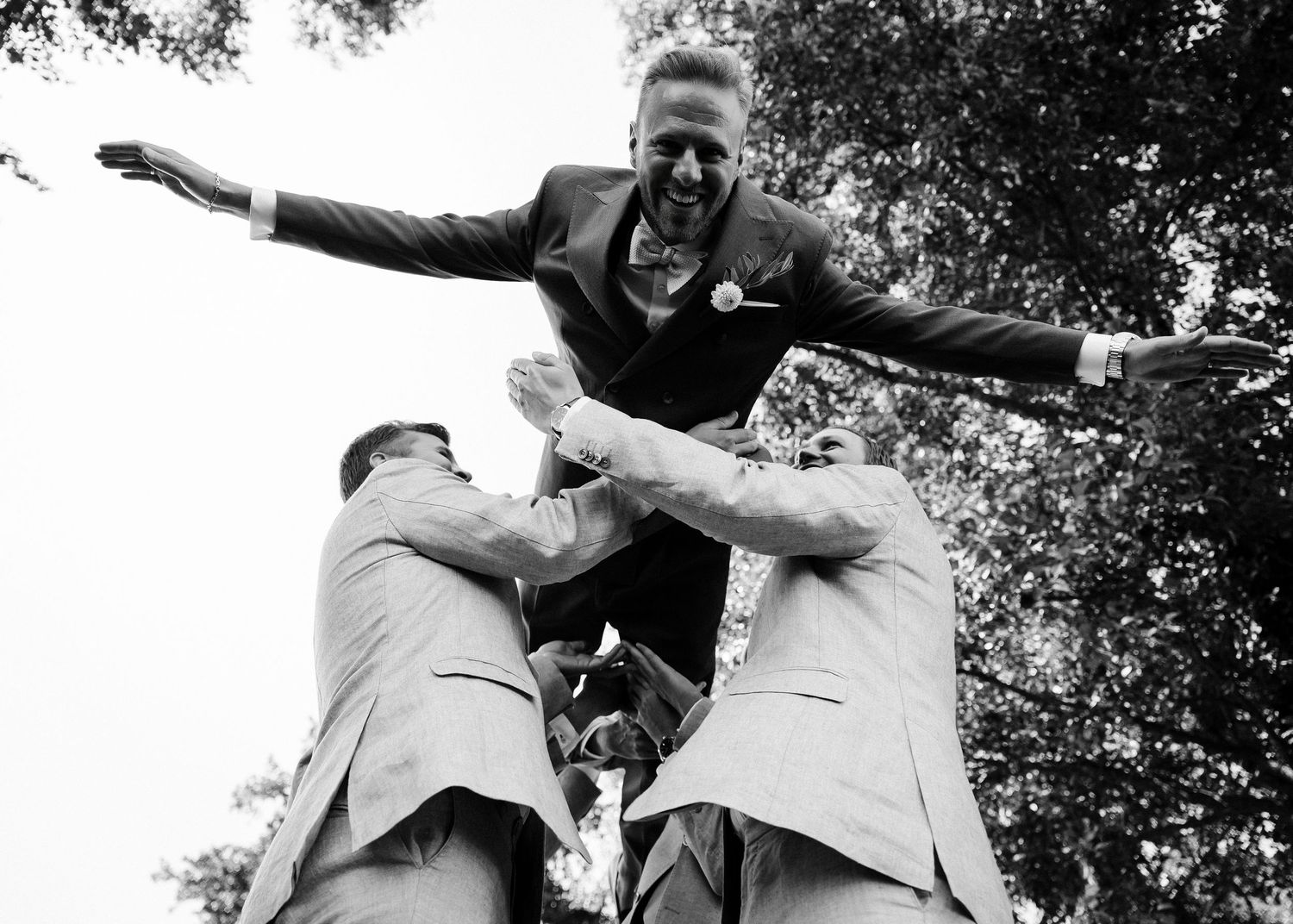 Black and white photo captures wedding party members jumping and posing playfully outdoors.