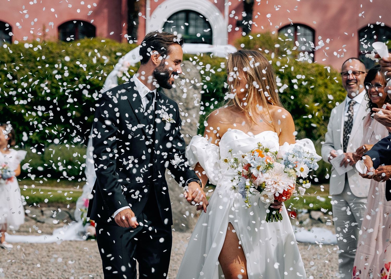 White confetti falling during outdoor wedding celebration with pink building in background.