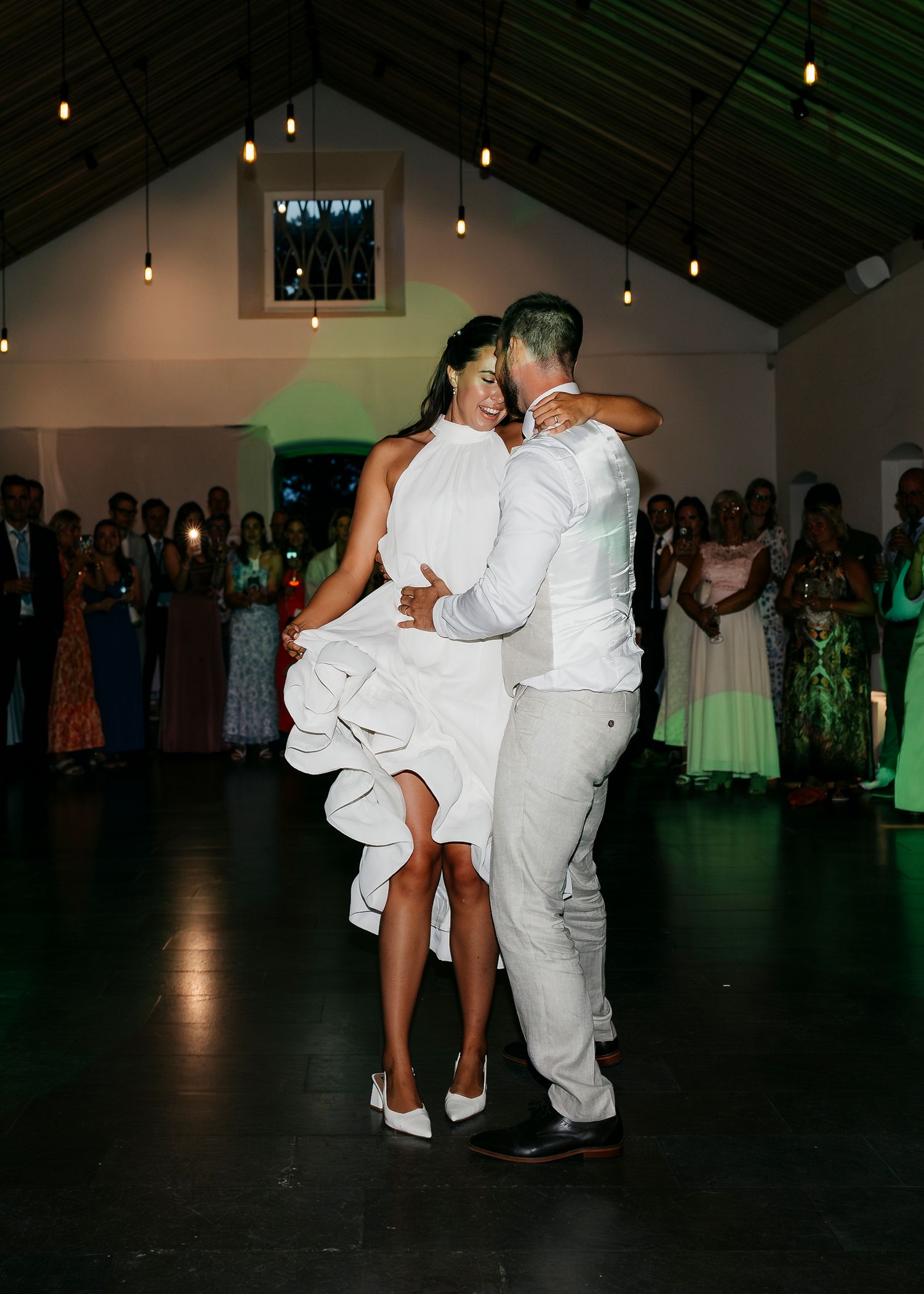 A couple dances intimately in an elegant venue with hanging lights and guests watching.