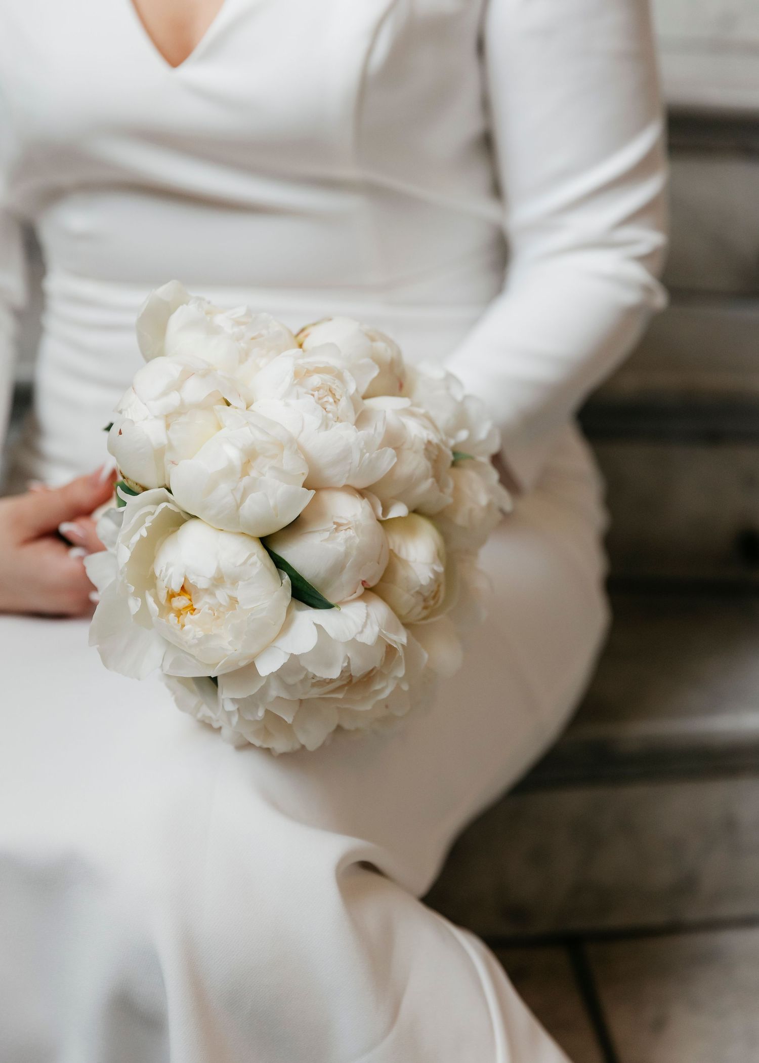 White bridal bouquet of fresh peonies held against a white dress.