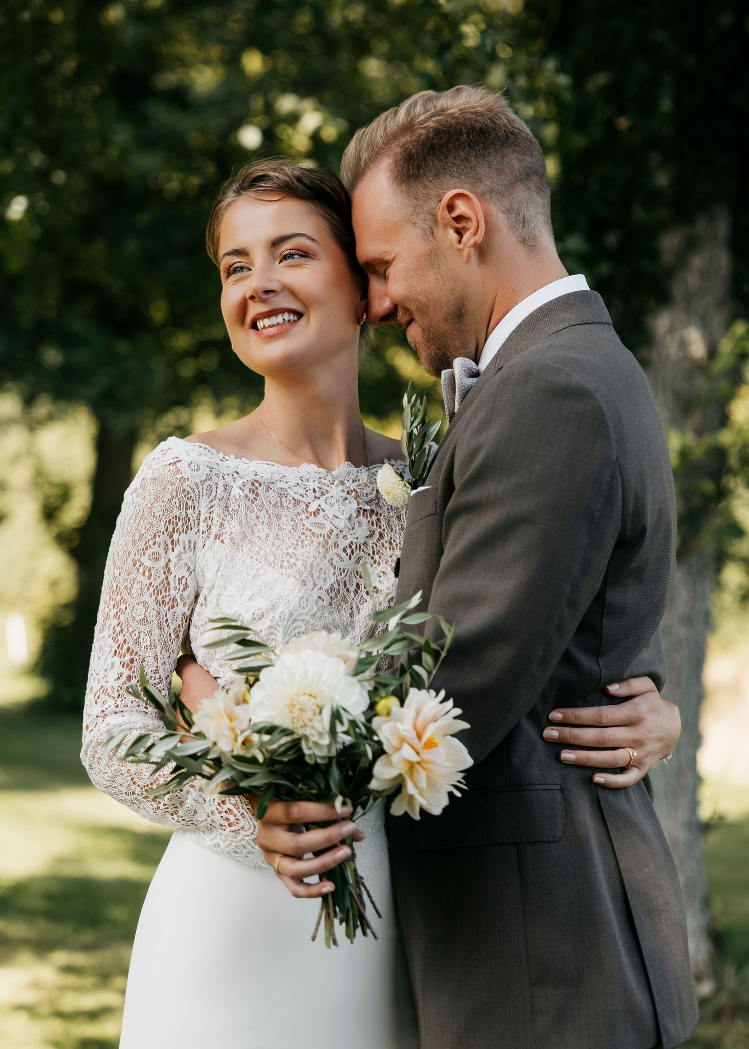 Newlyweds embrace while holding a white floral bouquet in a sunny garden setting.