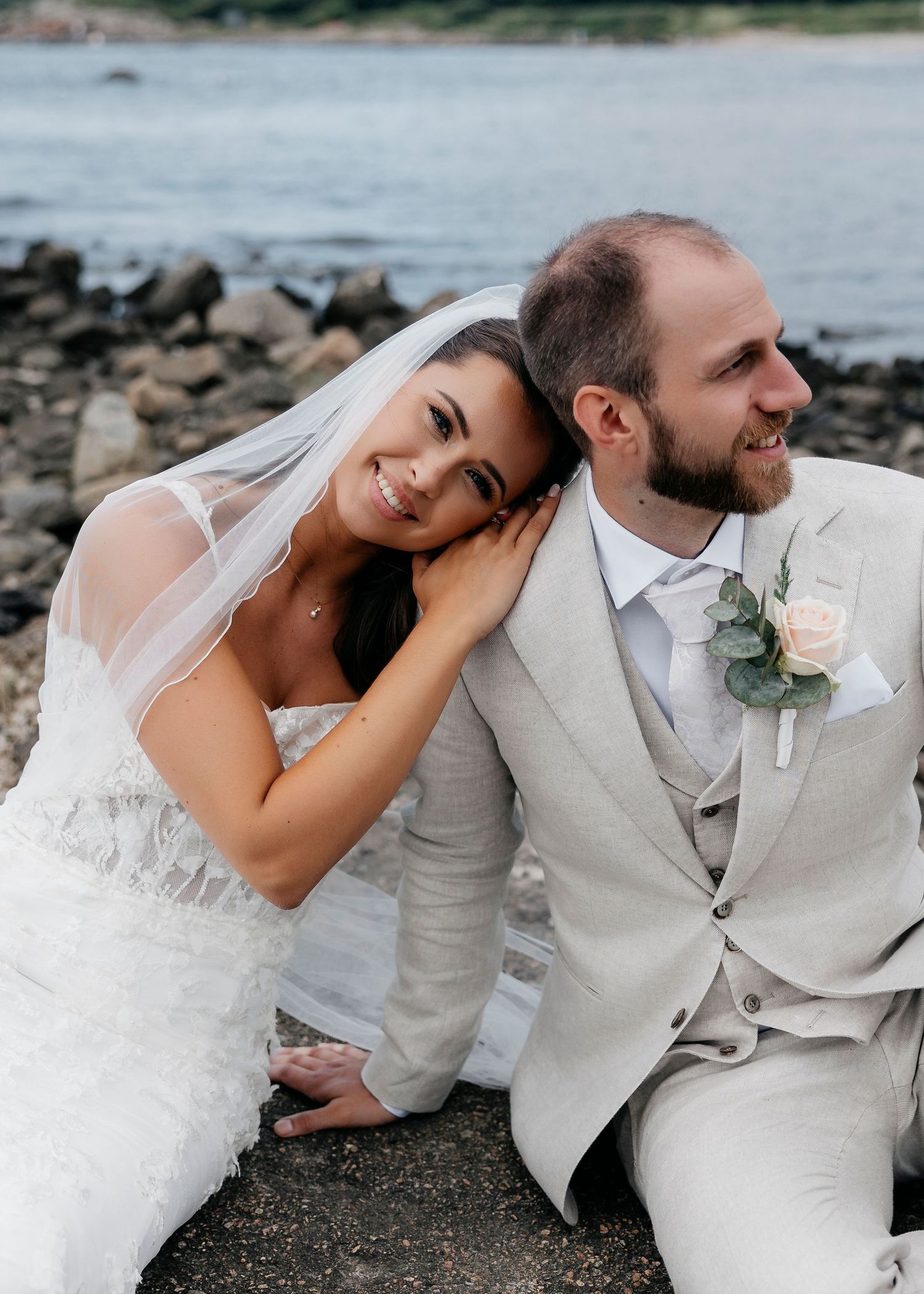 Romantic wedding couple sits on rocky beach shore sharing intimate moments by the water.