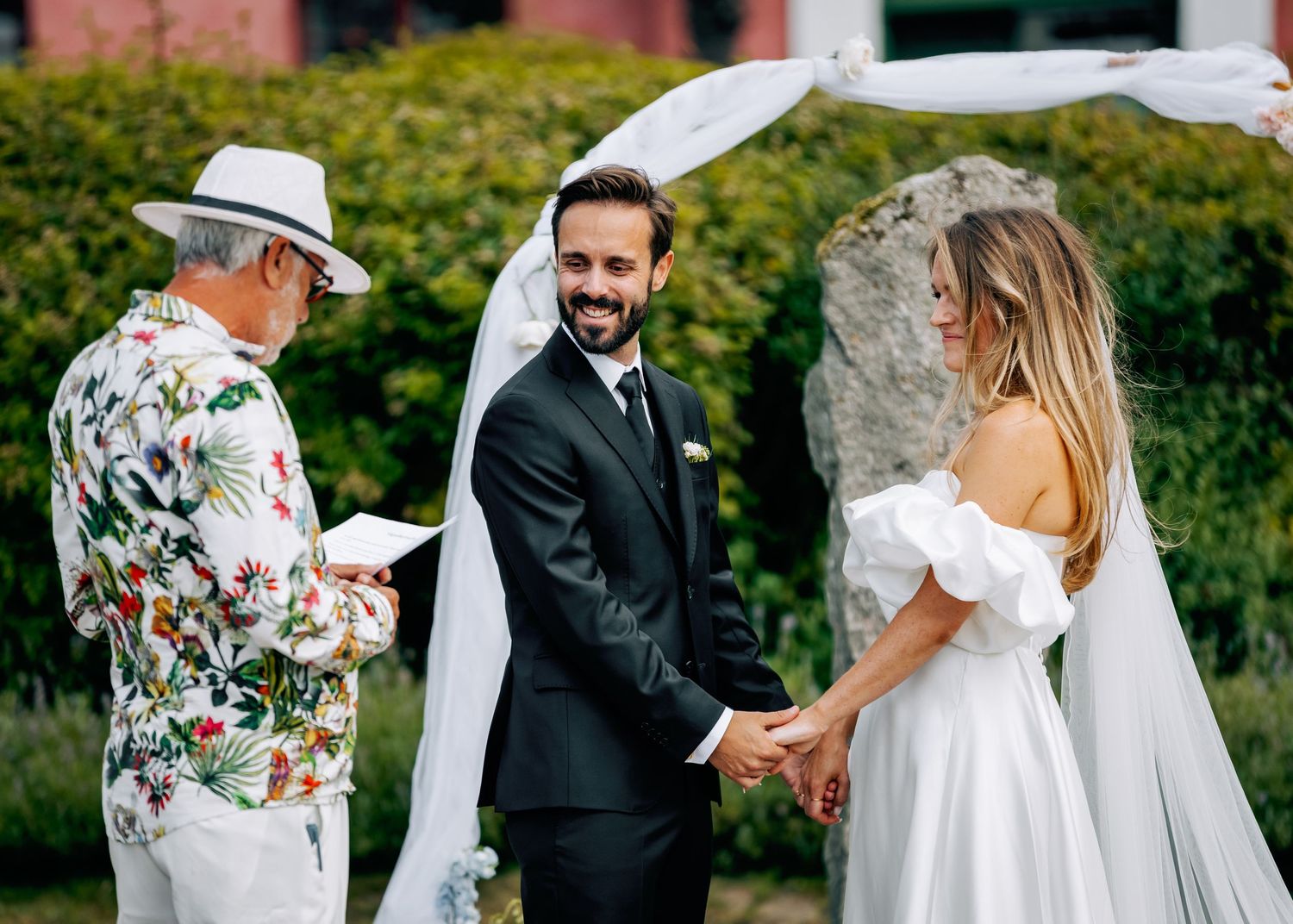 Wedding party celebrates outdoors with guests in floral attire and summer dresses.