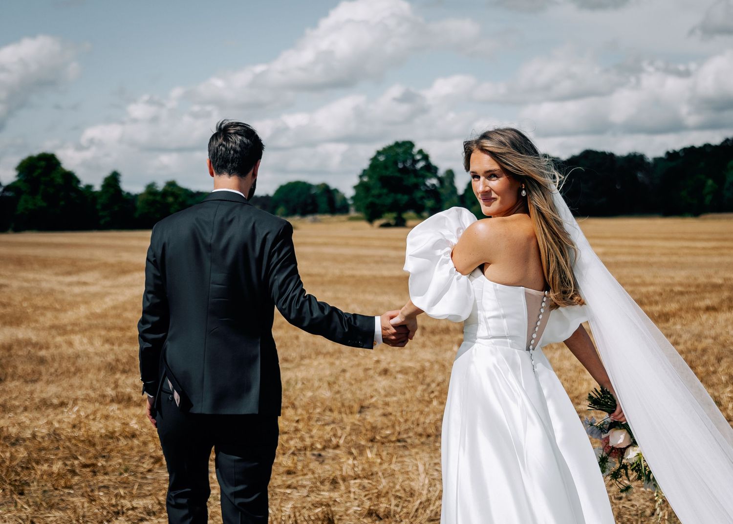 Wedding couple walking through golden field under cloudy summer sky.