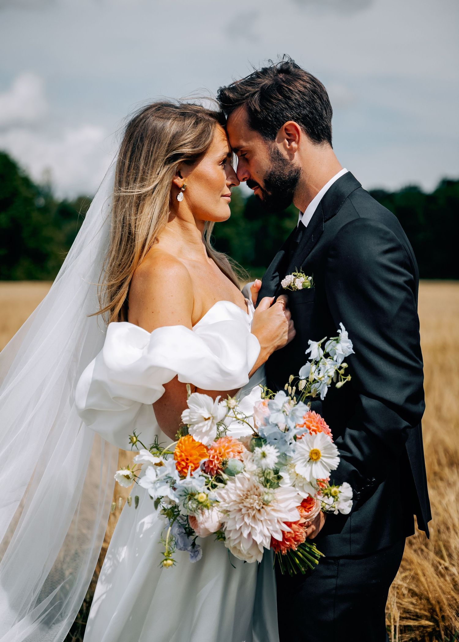 Newlywed couple shares intimate moment in wheat field with bride holding vibrant summer bouquet of mixed flowers.