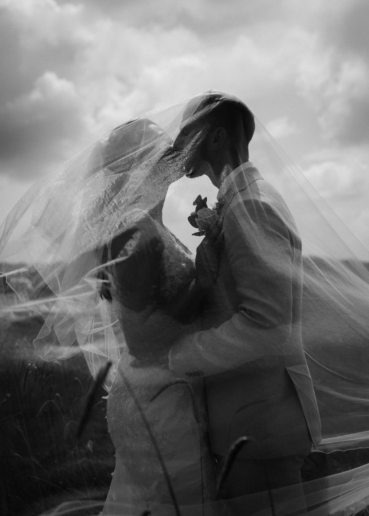 Dramatic black and white shot of couple with flowing veil against cloudy sky.