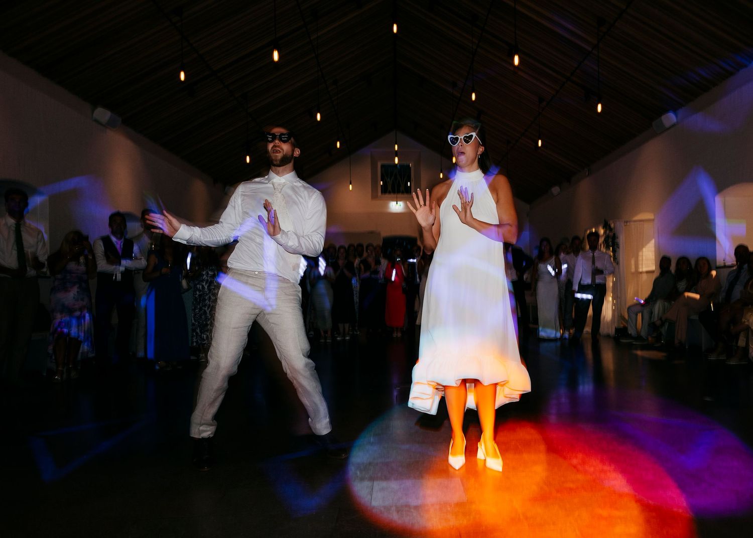 Wedding couple performs an energetic dance routine in a reception hall with colorful lighting.