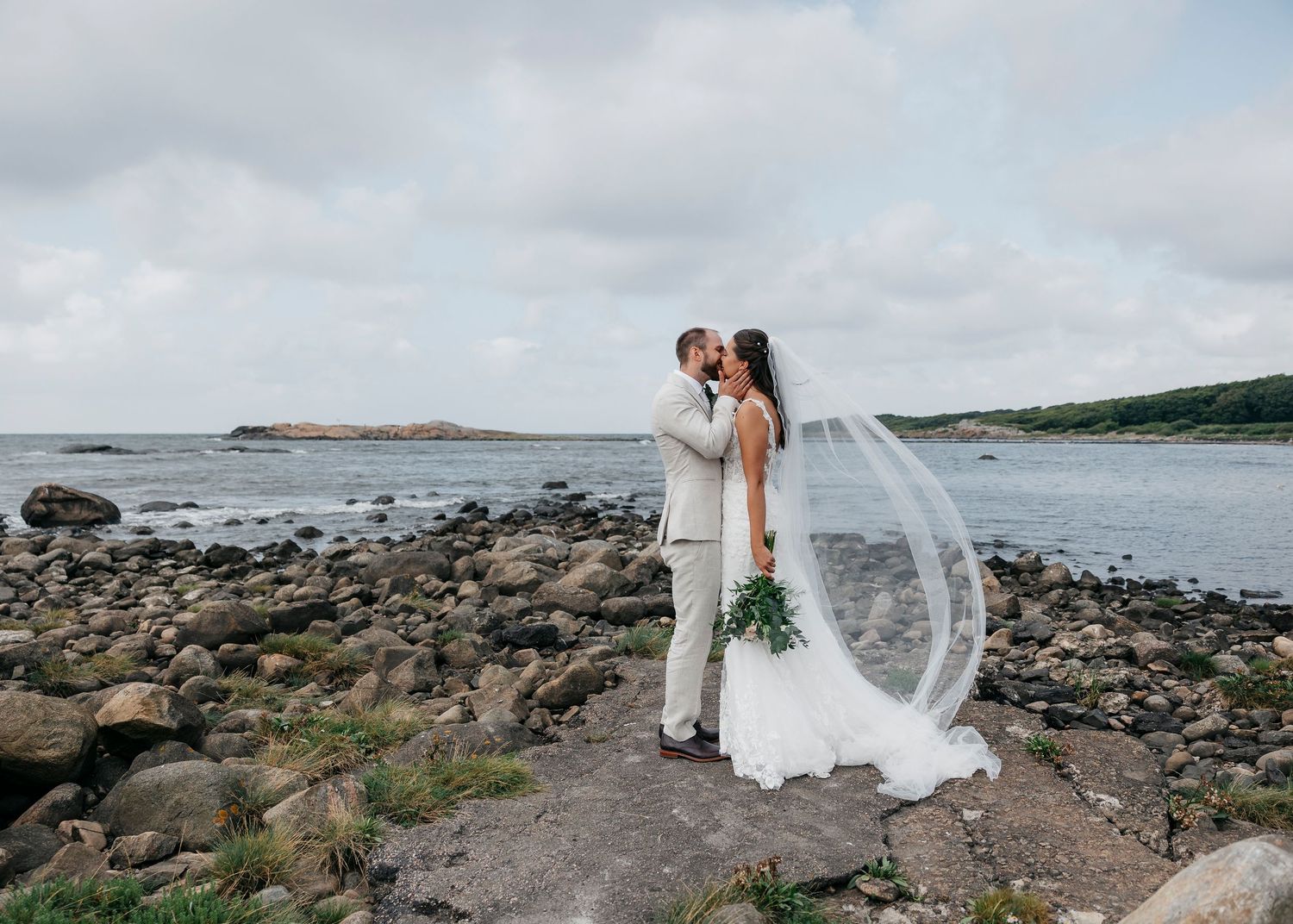 Couple in wedding attire poses on rocky coastline with ocean views.