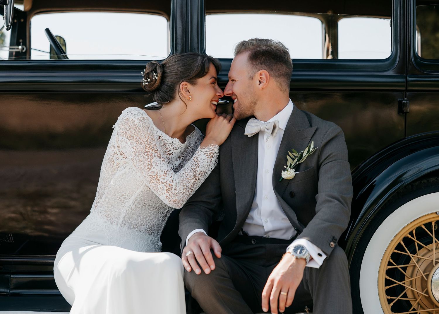Newlyweds pose romantically against classic black automobile.